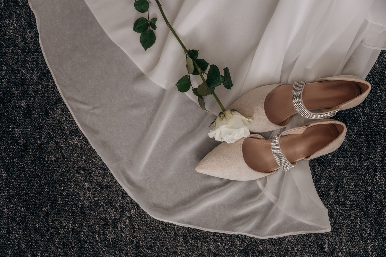 Elegant bridal flat shoes with crystal strap beside a white rose on flowing wedding dress fabric.