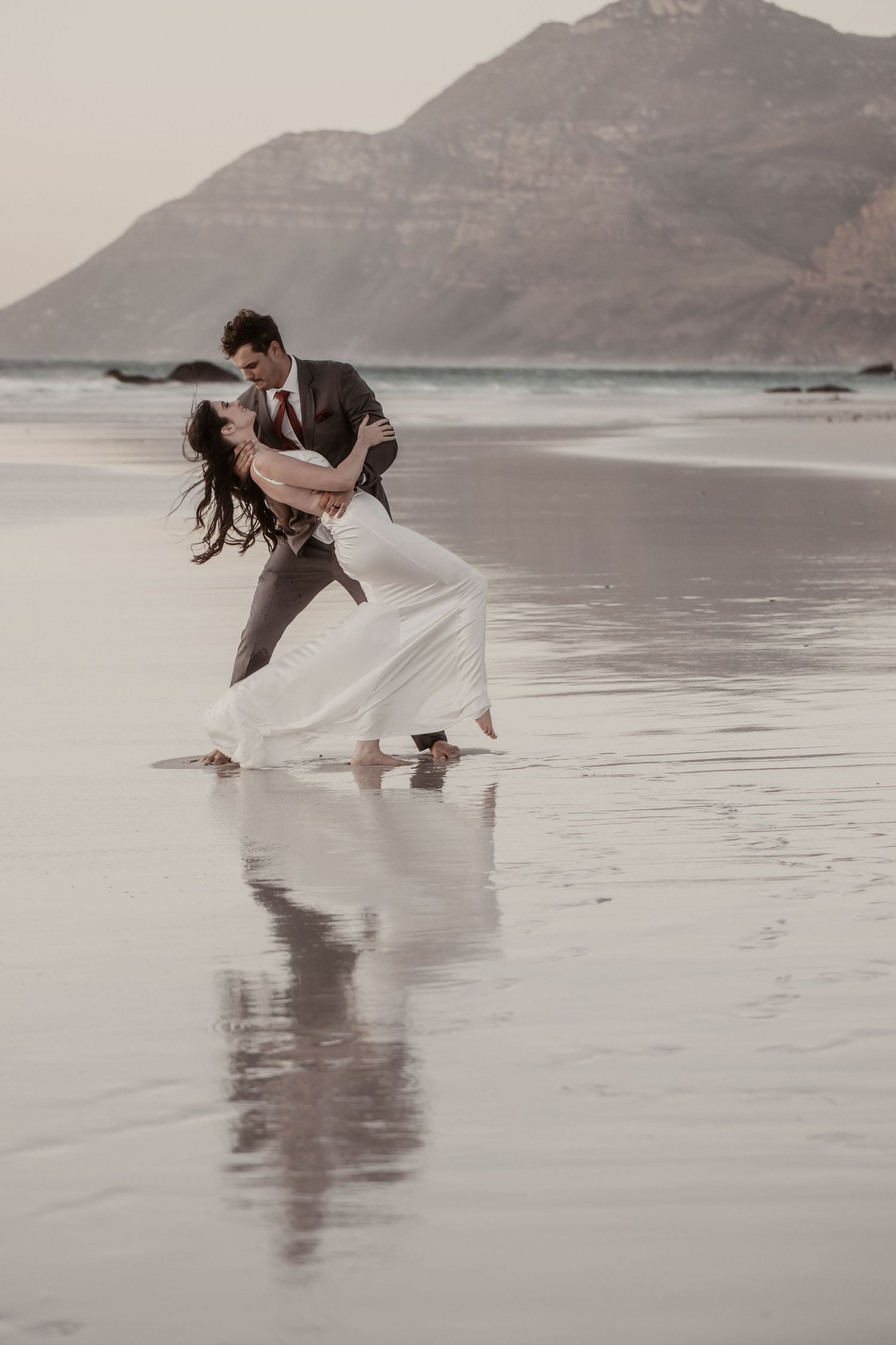 A couple in wedding attire shares a romantic dip kiss on a reflective beach with a mountain backdrop.