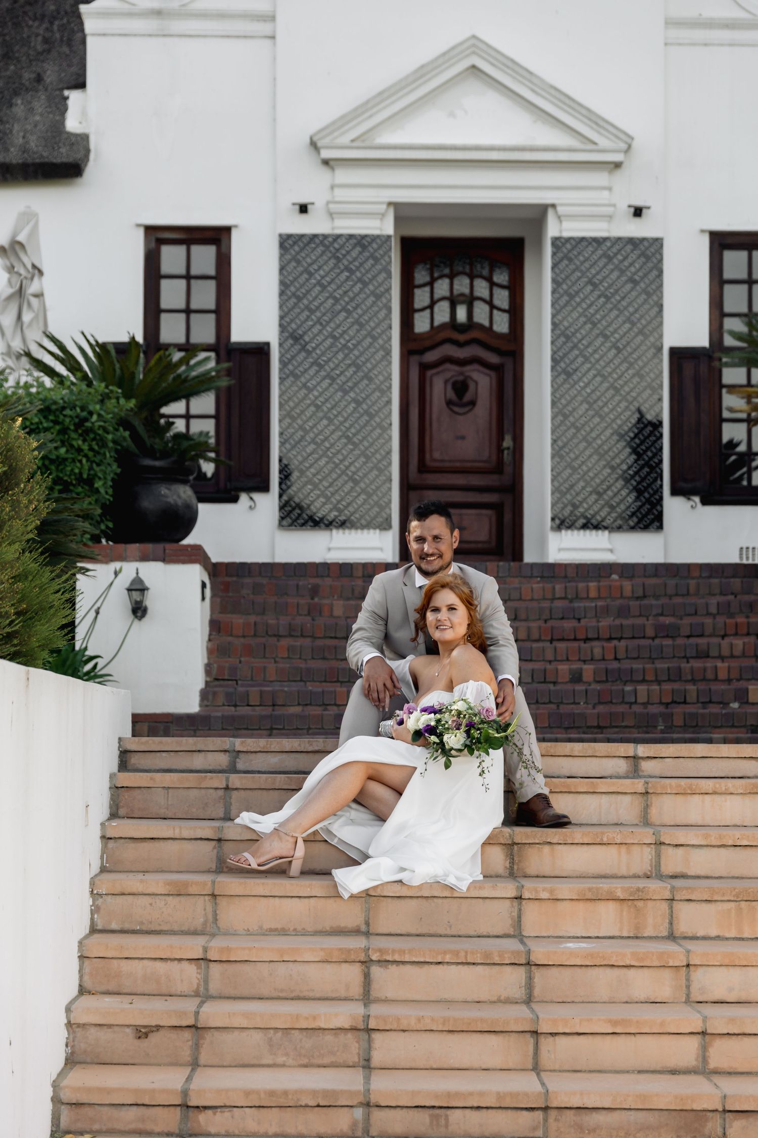 Bride and groom sitting on stone steps of elegant white building, bride holding bouquet in white dress.