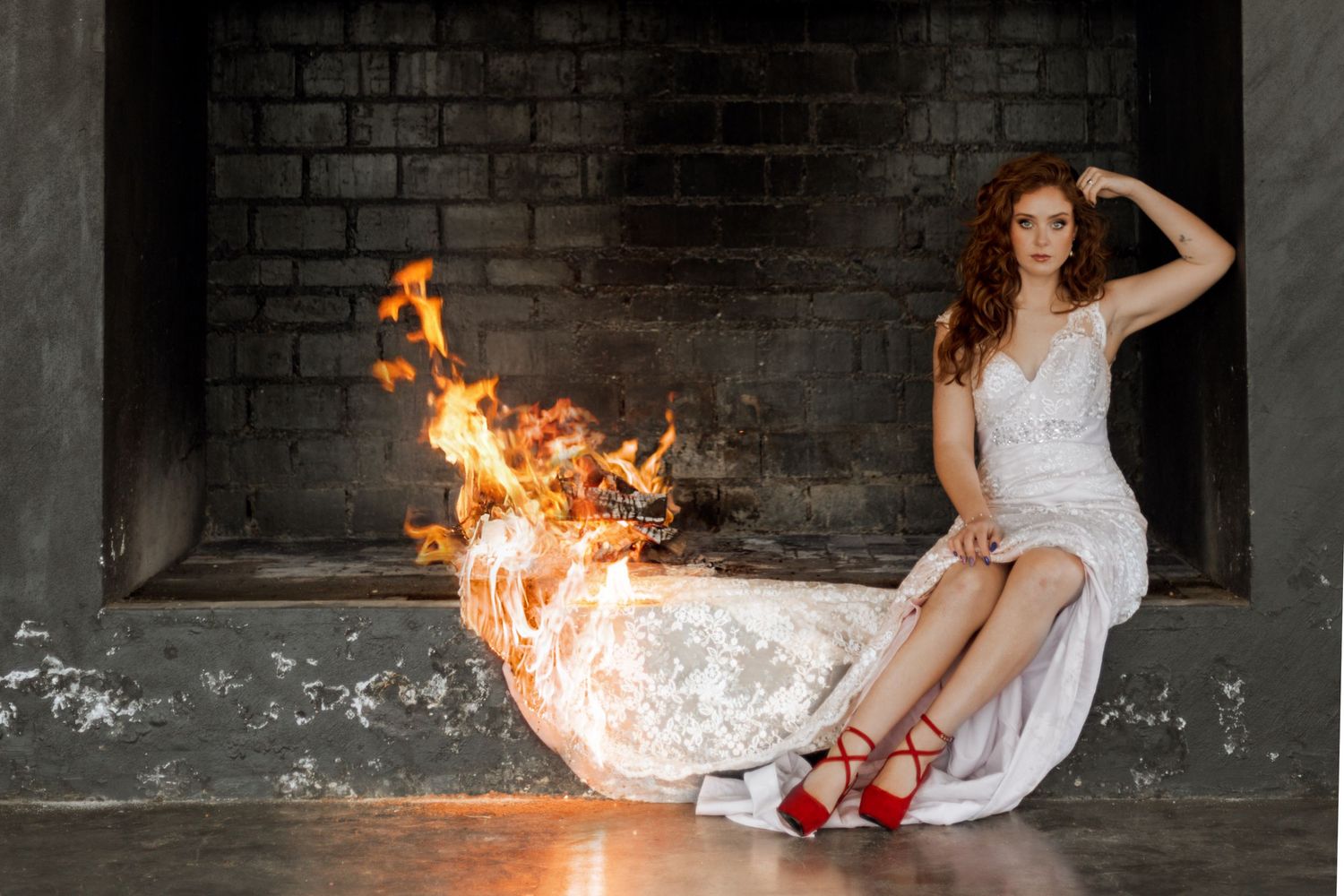 A figure in a flowing white dress reclines on the floor near dramatic flames against a dark brick wall.