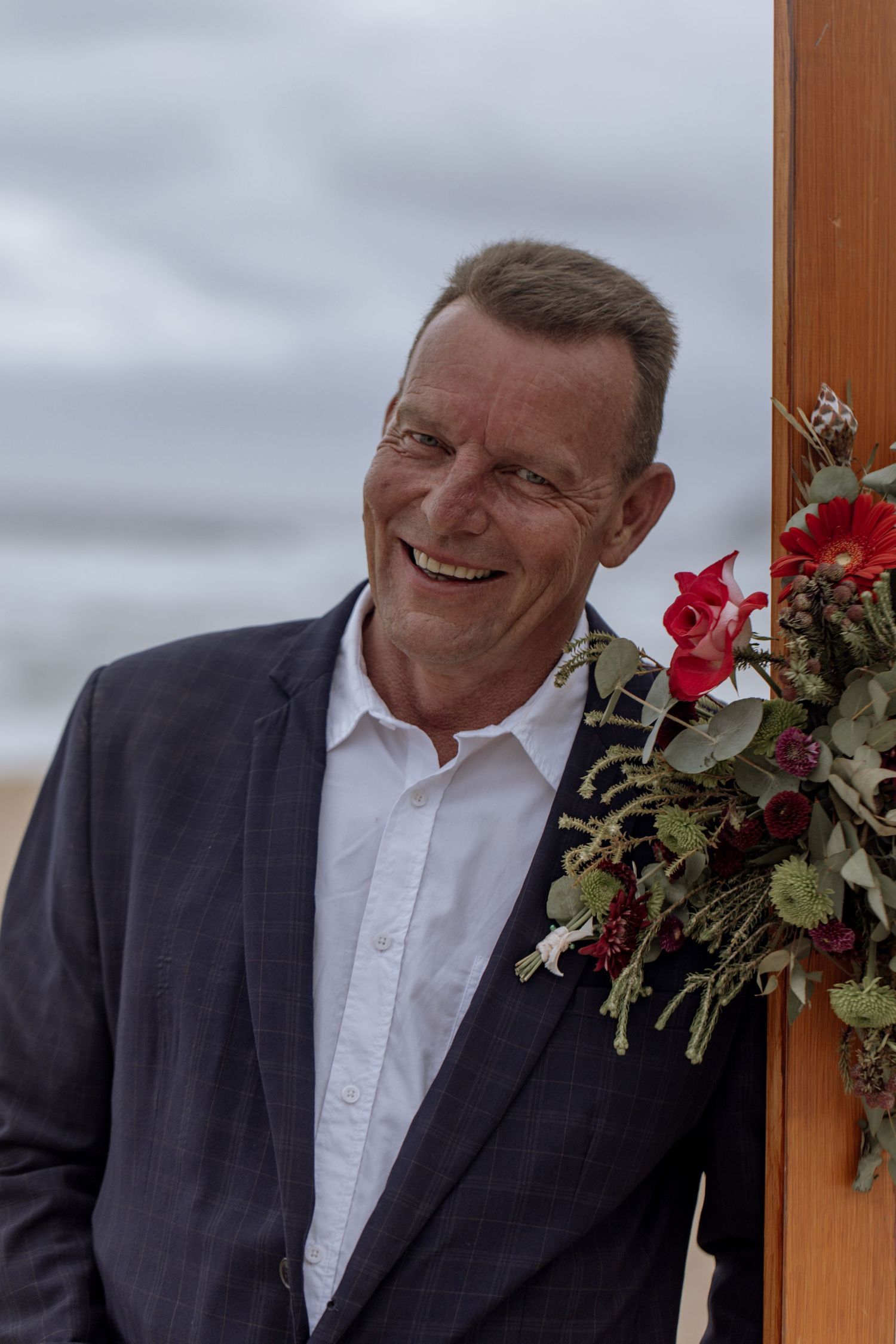 Smiling person in dark suit and white shirt stands next to decorative red floral arrangement against cloudy sky.