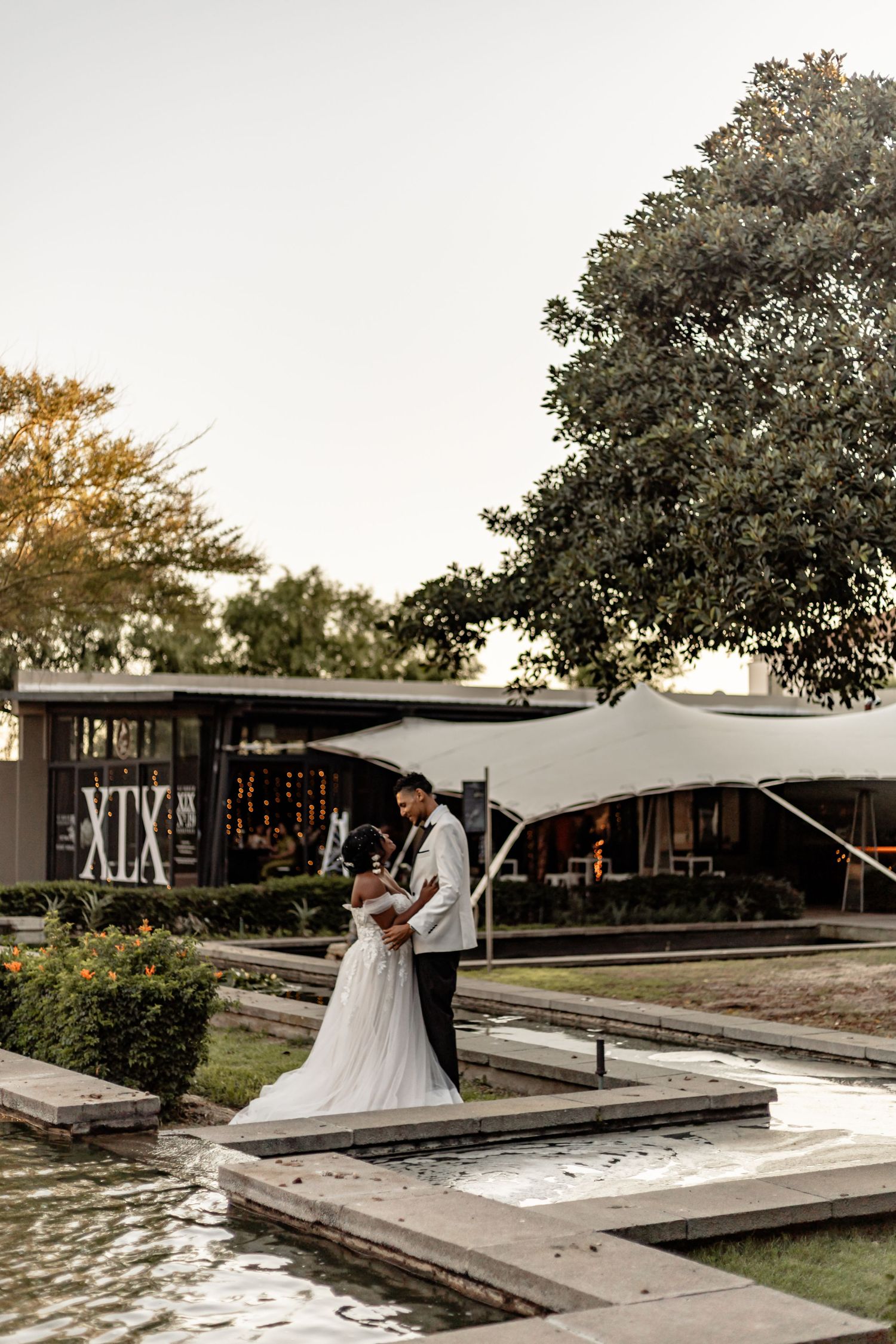 Wedding couple embracing by modern venue with water feature at sunset.