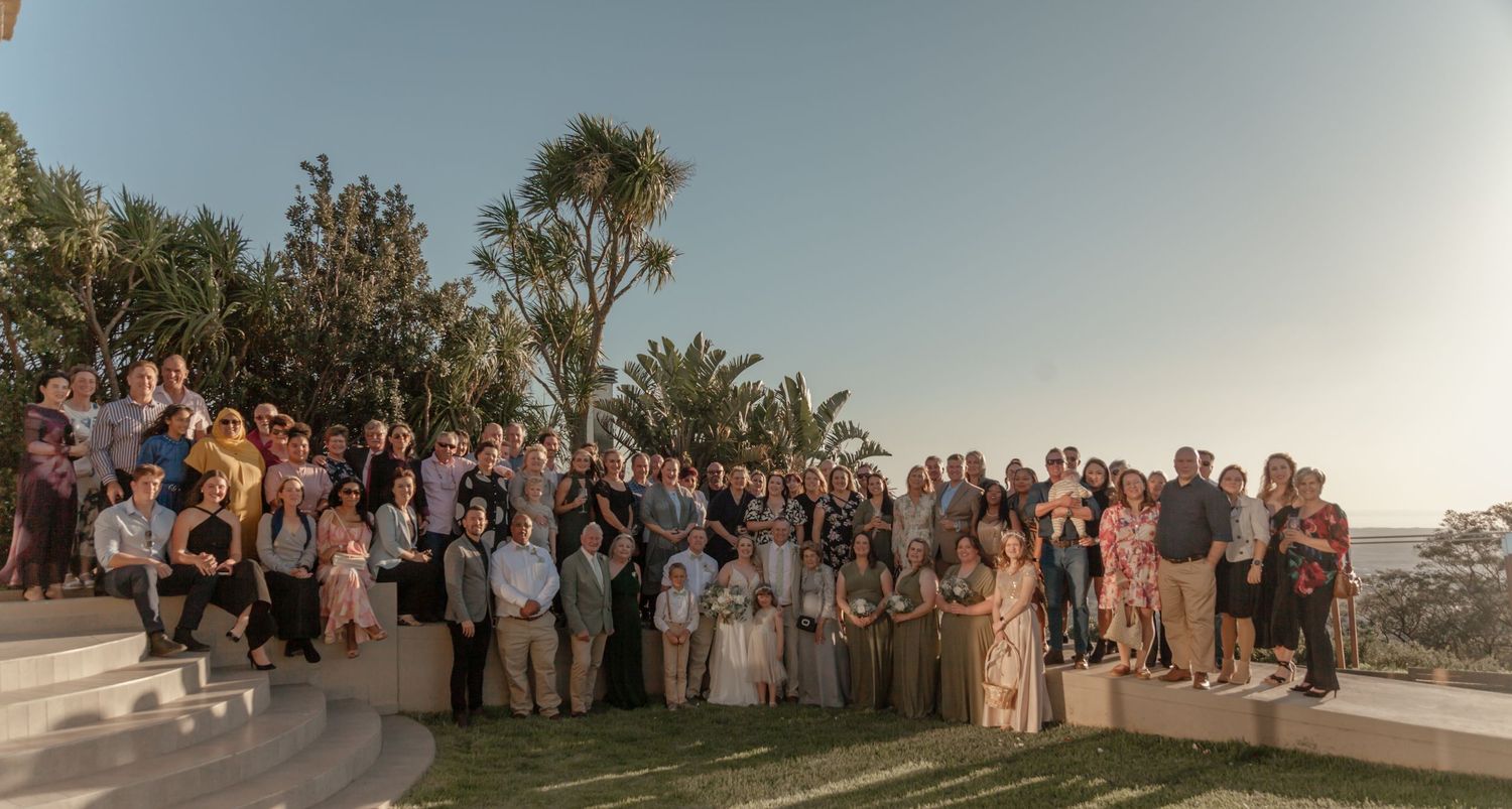 Large group of wedding guests gathered outdoors at sunset for a scenic group photo on a warm Mediterranean evening.