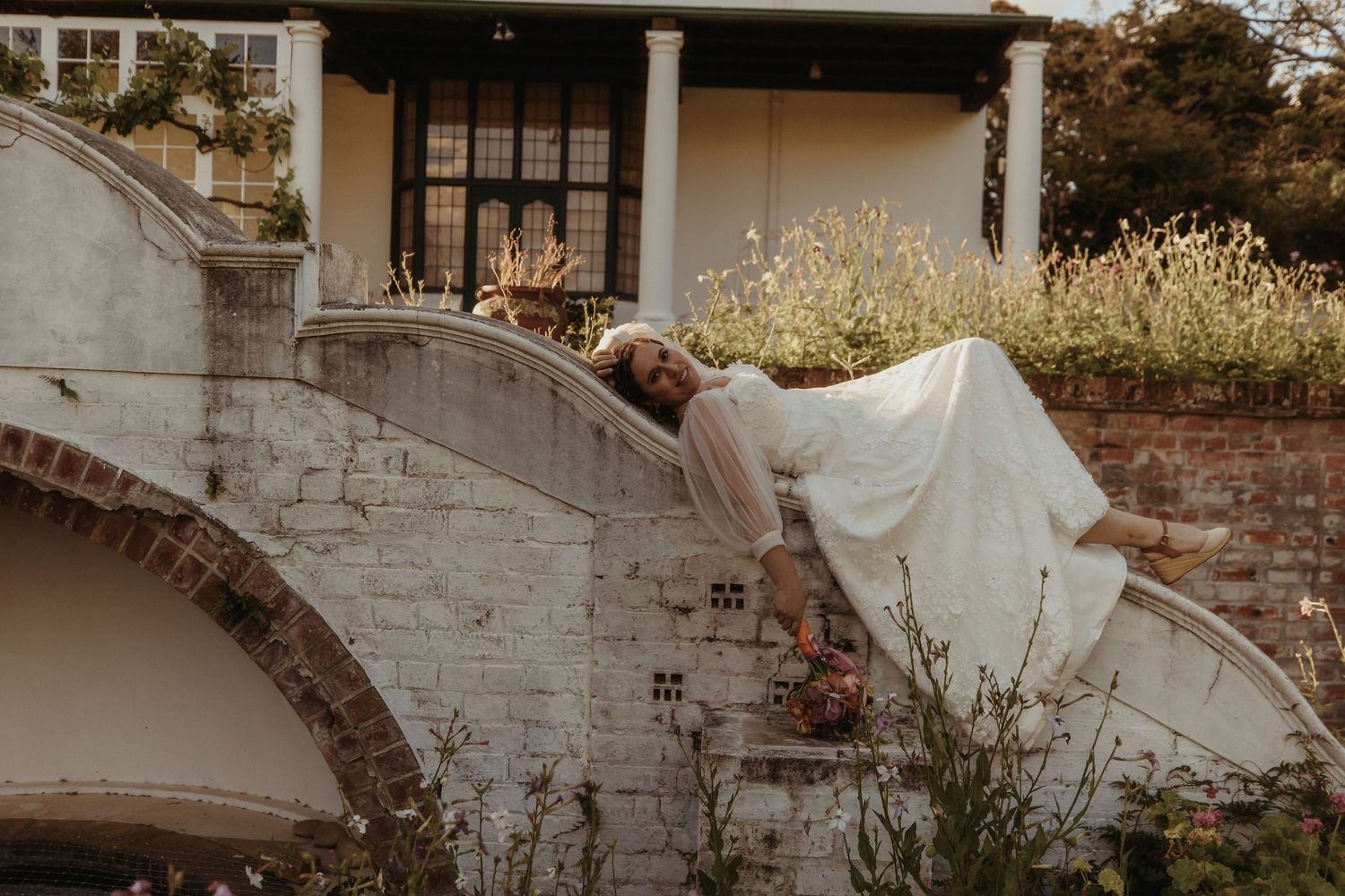 A couple in formal wedding attire embraces while standing against a rustic stone wall in warm sunlight.
