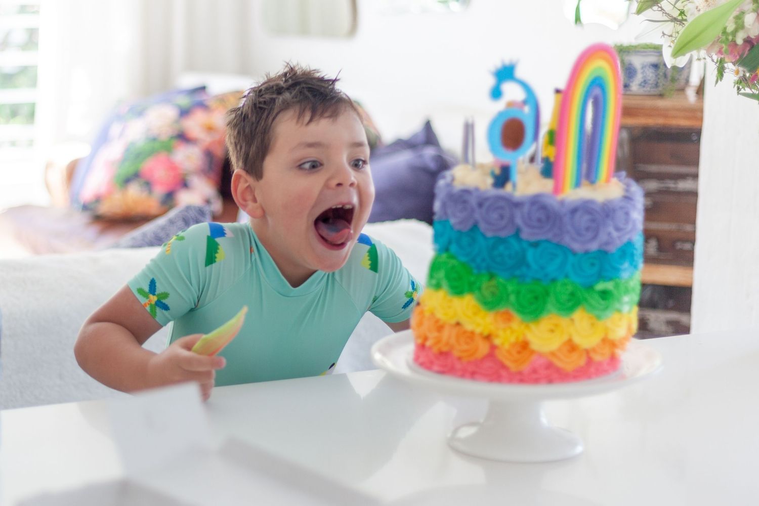 Excited young boy with open mouth looking at a colorful rainbow birthday cake with number candles on white table.