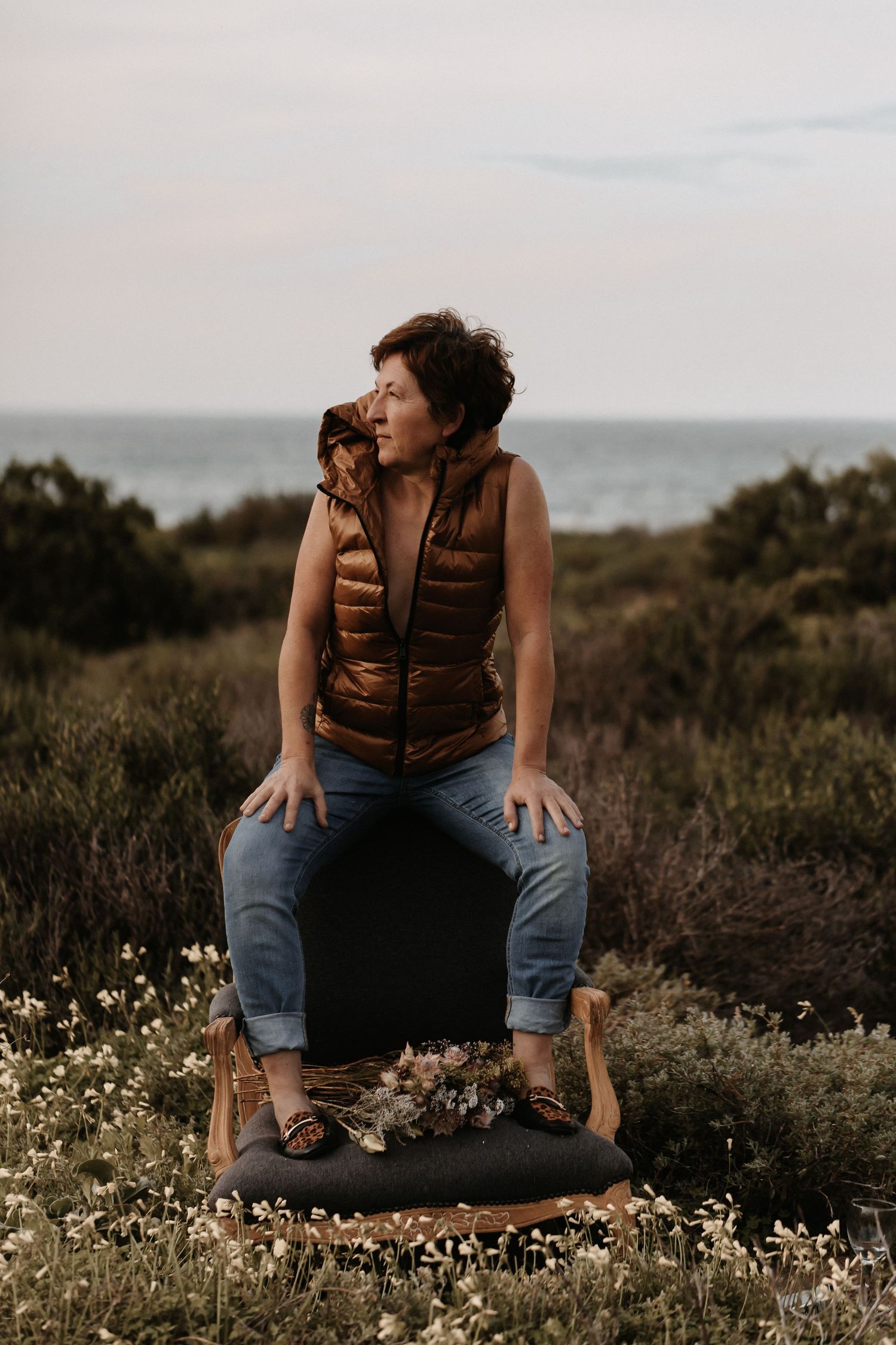 Person in brown puffer vest and jeans sits on a black stool against coastal landscape with wildflowers at sunset.
