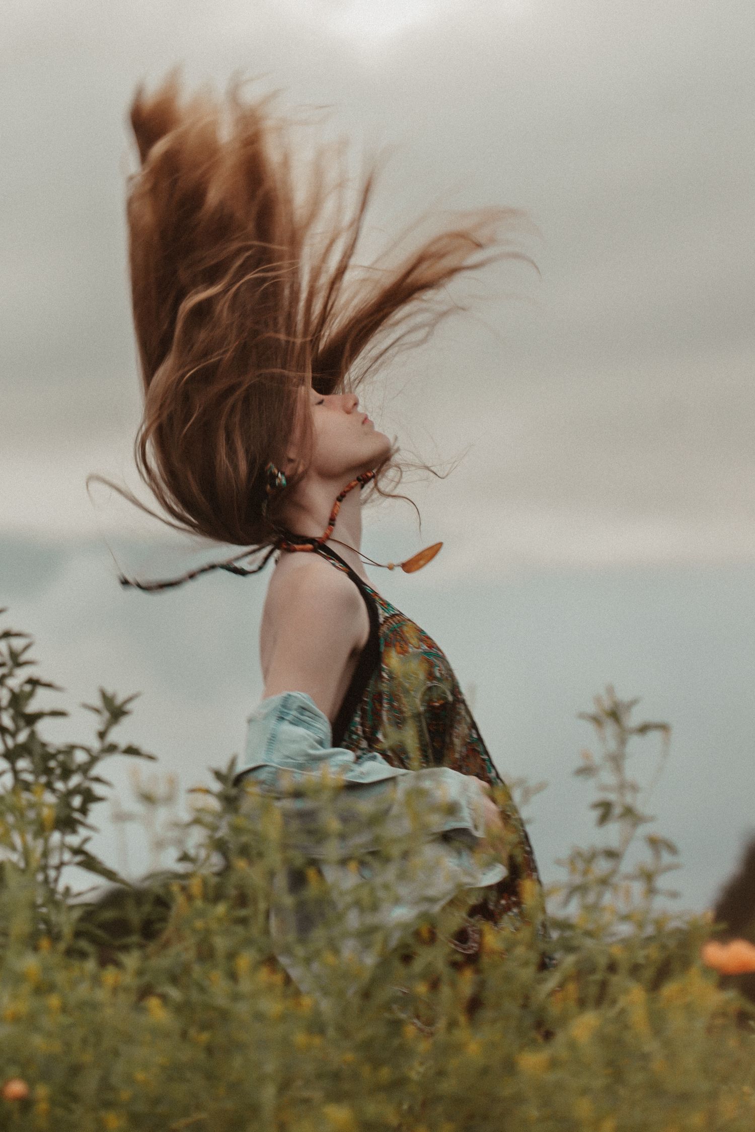 Long auburn hair whips through the air as someone spins among wildflowers against a cloudy sky.