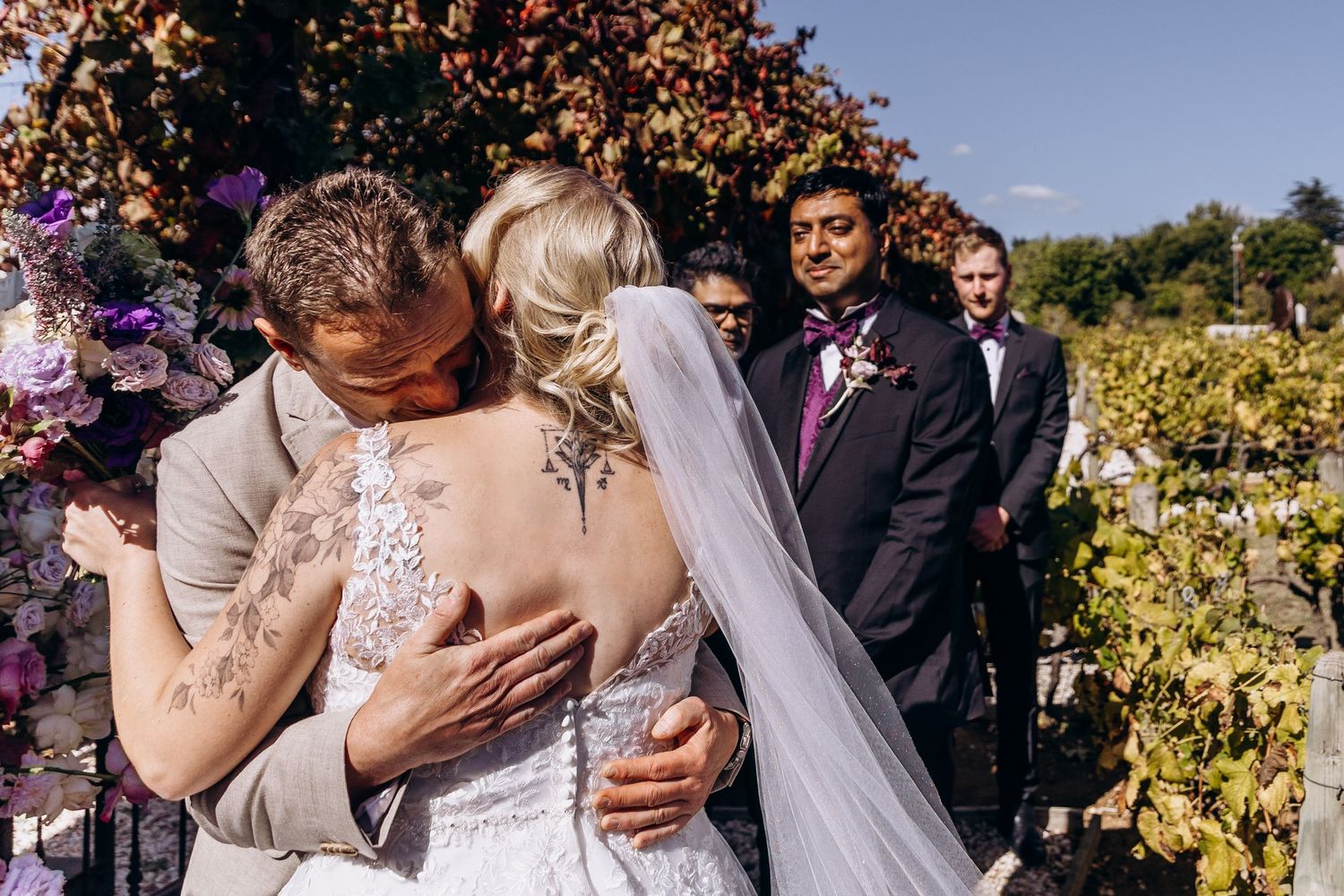 Bride and groom share first kiss at outdoor vineyard wedding ceremony with groomsmen watching in background.