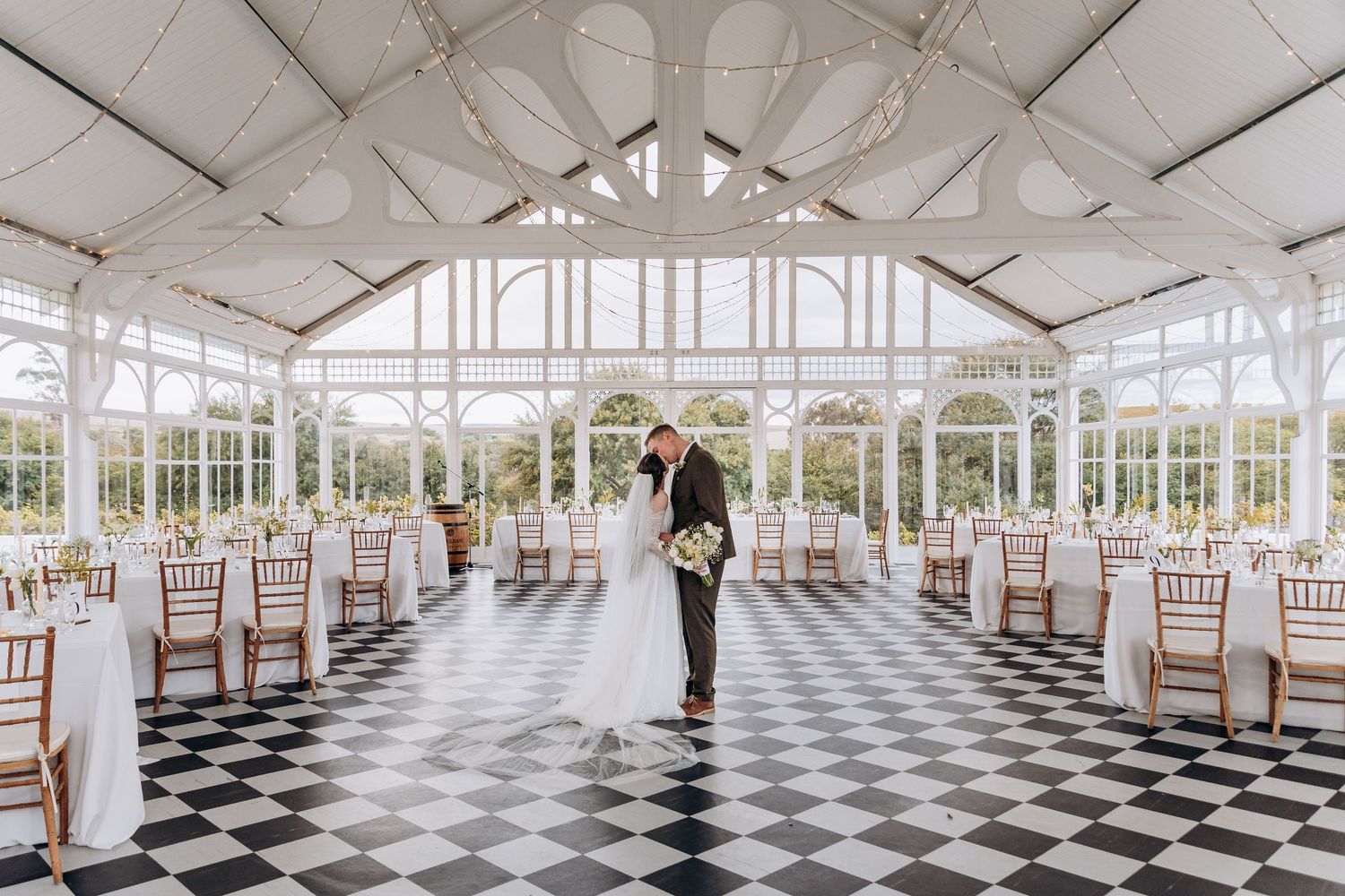 Bride and groom share a romantic kiss in an elegant glass conservatory wedding venue with checkered floors and white-draped tables.