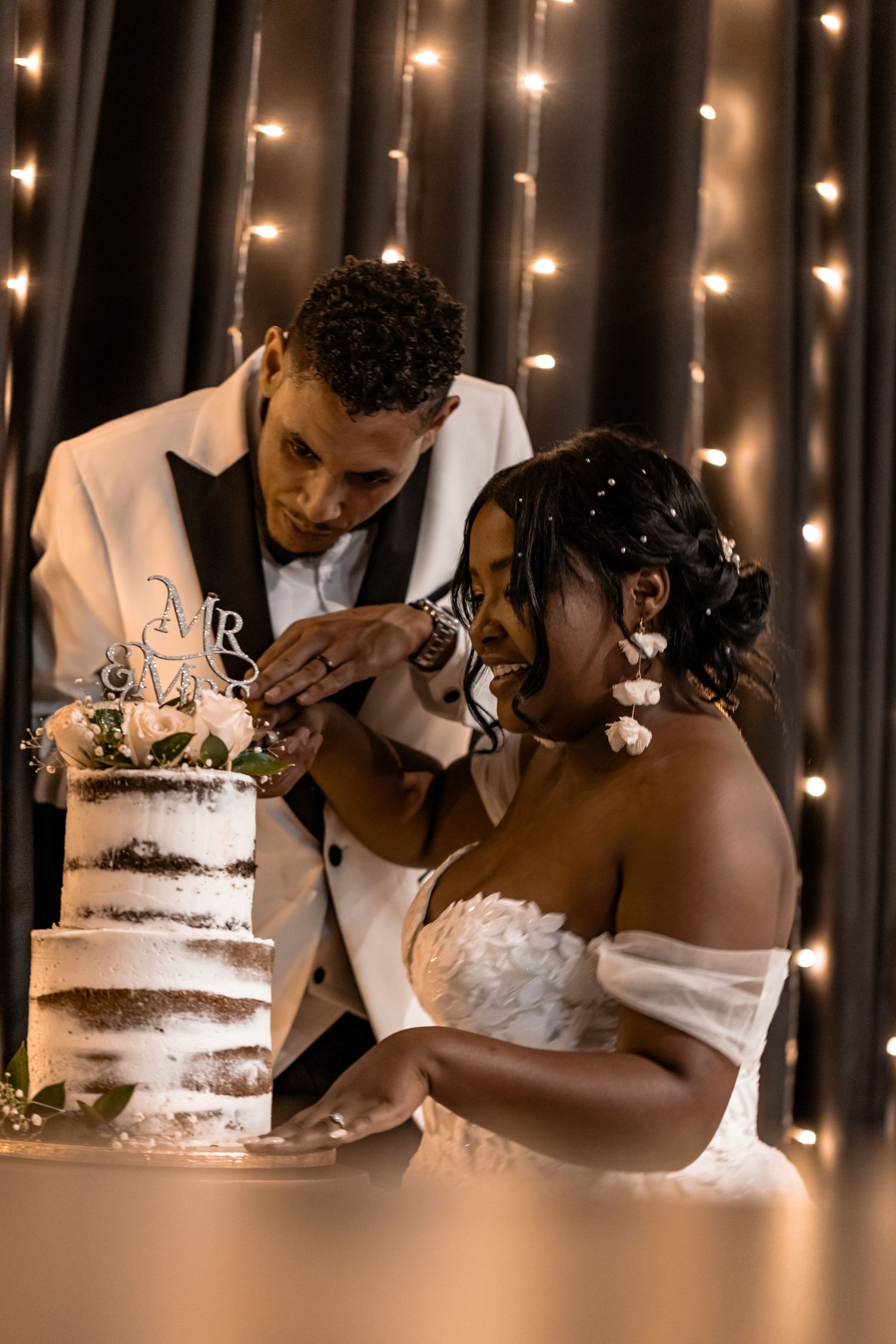 Newlyweds share a sweet moment while cutting their rustic semi-naked wedding cake in front of twinkling lights.