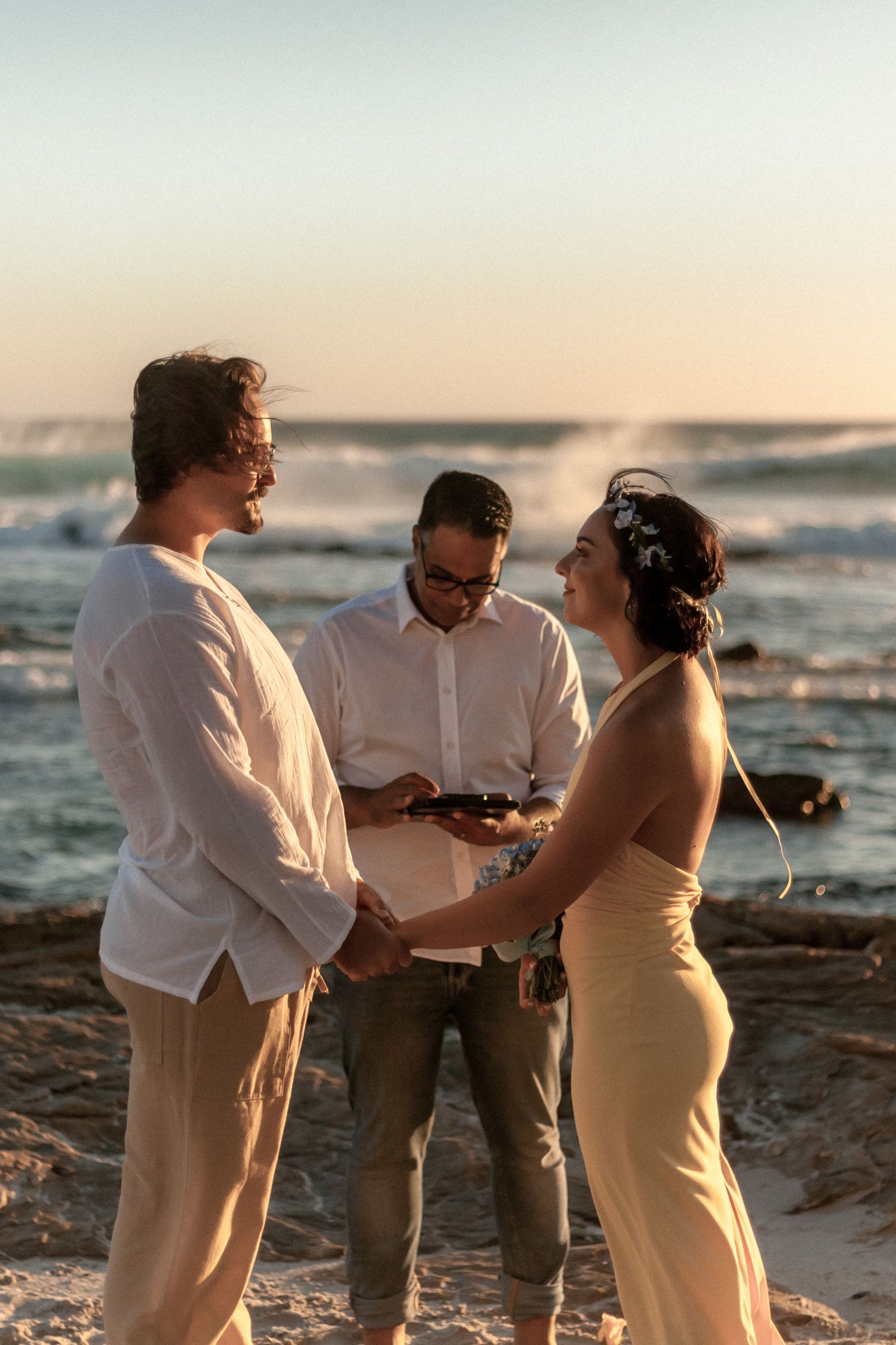 A romantic sunset beach wedding ceremony with three people standing by the ocean's edge at golden hour.