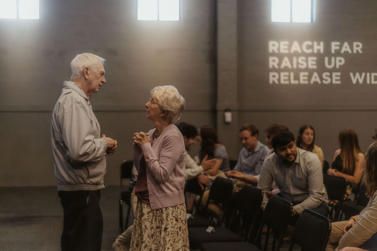 Elderly couple dances together in a dimly lit room while others watch from tables in the background.
