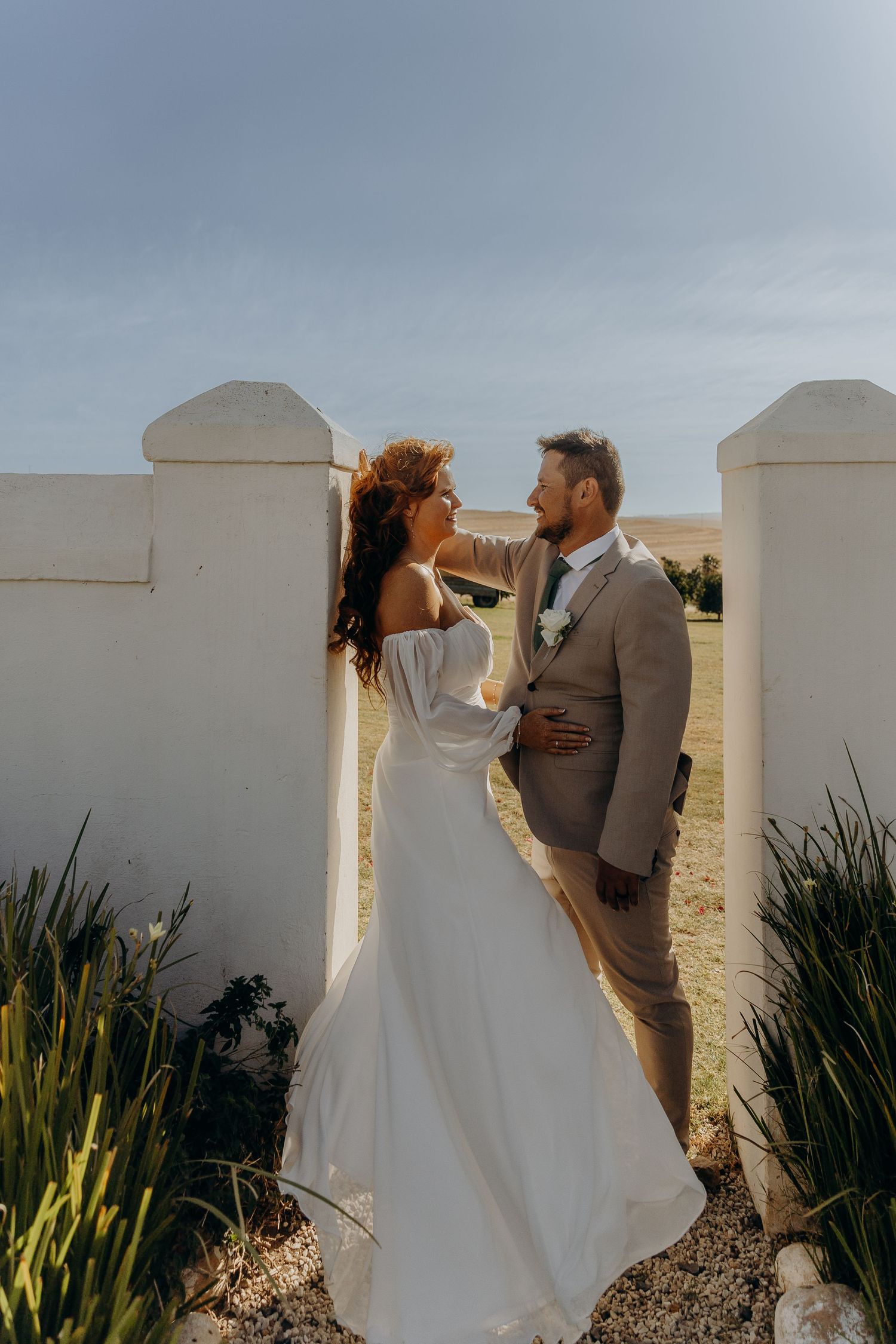 Bride and groom share a romantic moment between white stone pillars at an outdoor wedding venue.