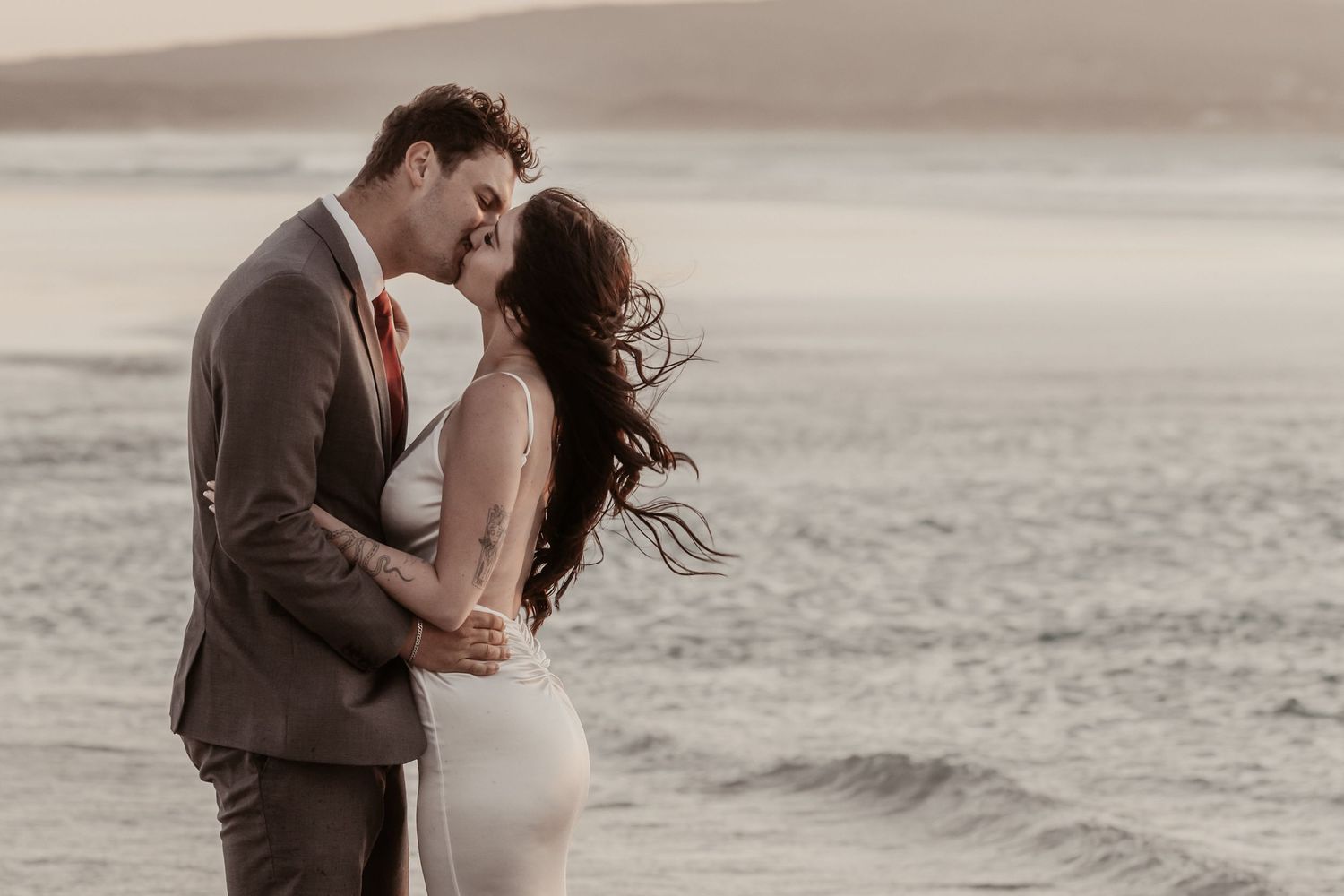 A couple shares a romantic kiss on a beach at sunset, with the bride in a white gown and groom in a gray suit.