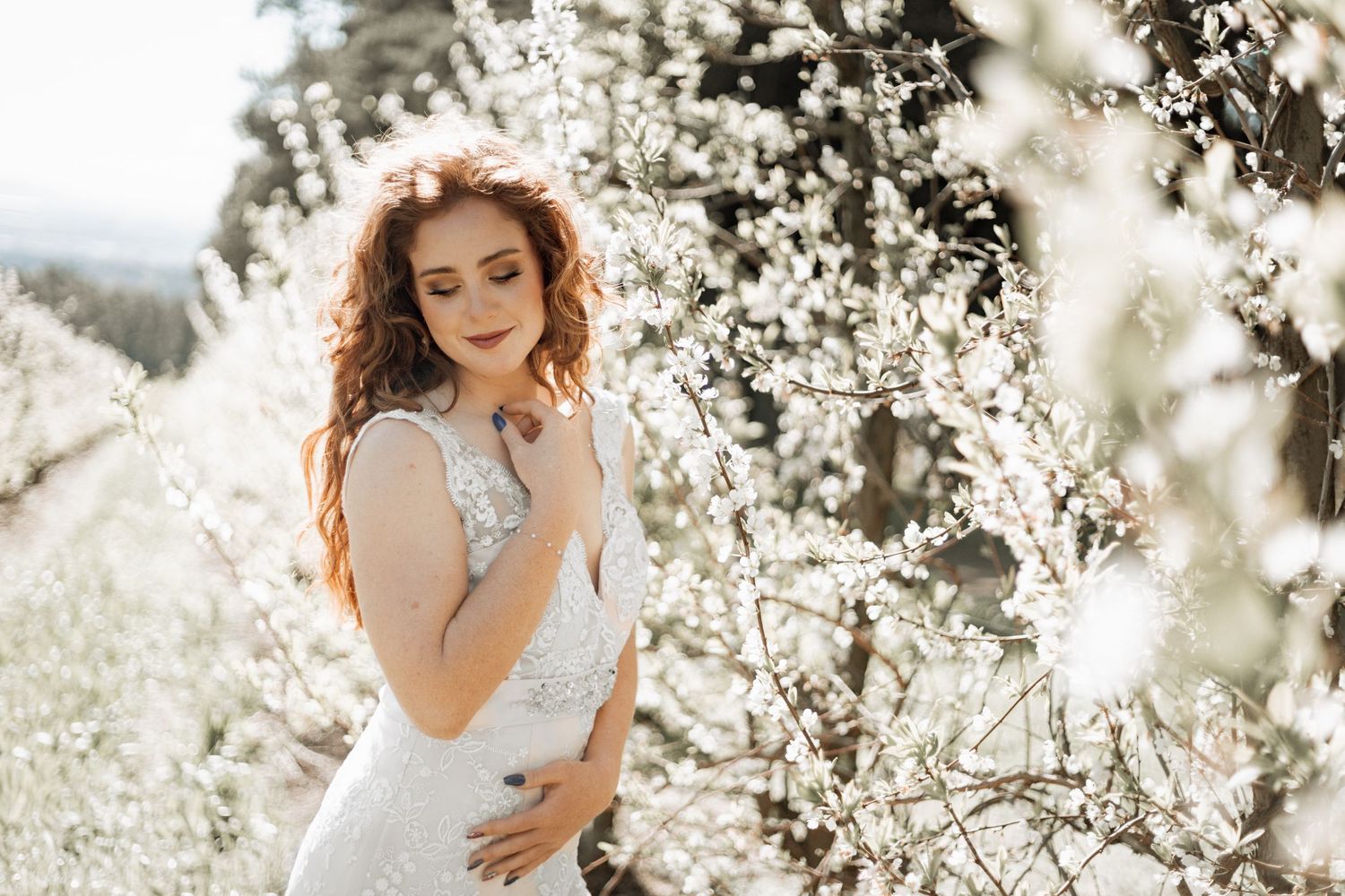 Woman in white flowing dress stands gracefully among delicate cherry blossoms on a spring day.