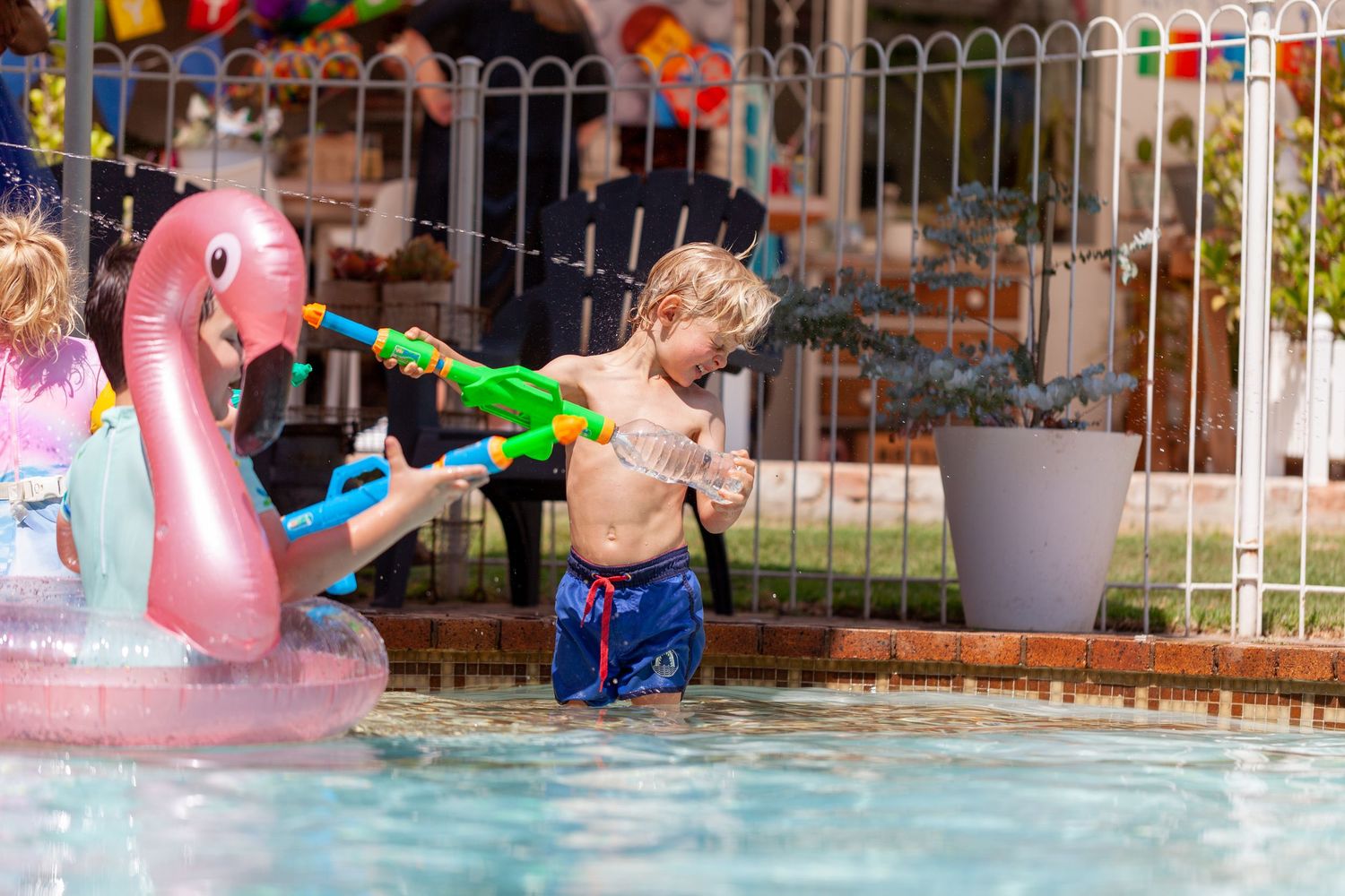Child playing with a water gun in a pool beside a pink flamingo float on a sunny day.