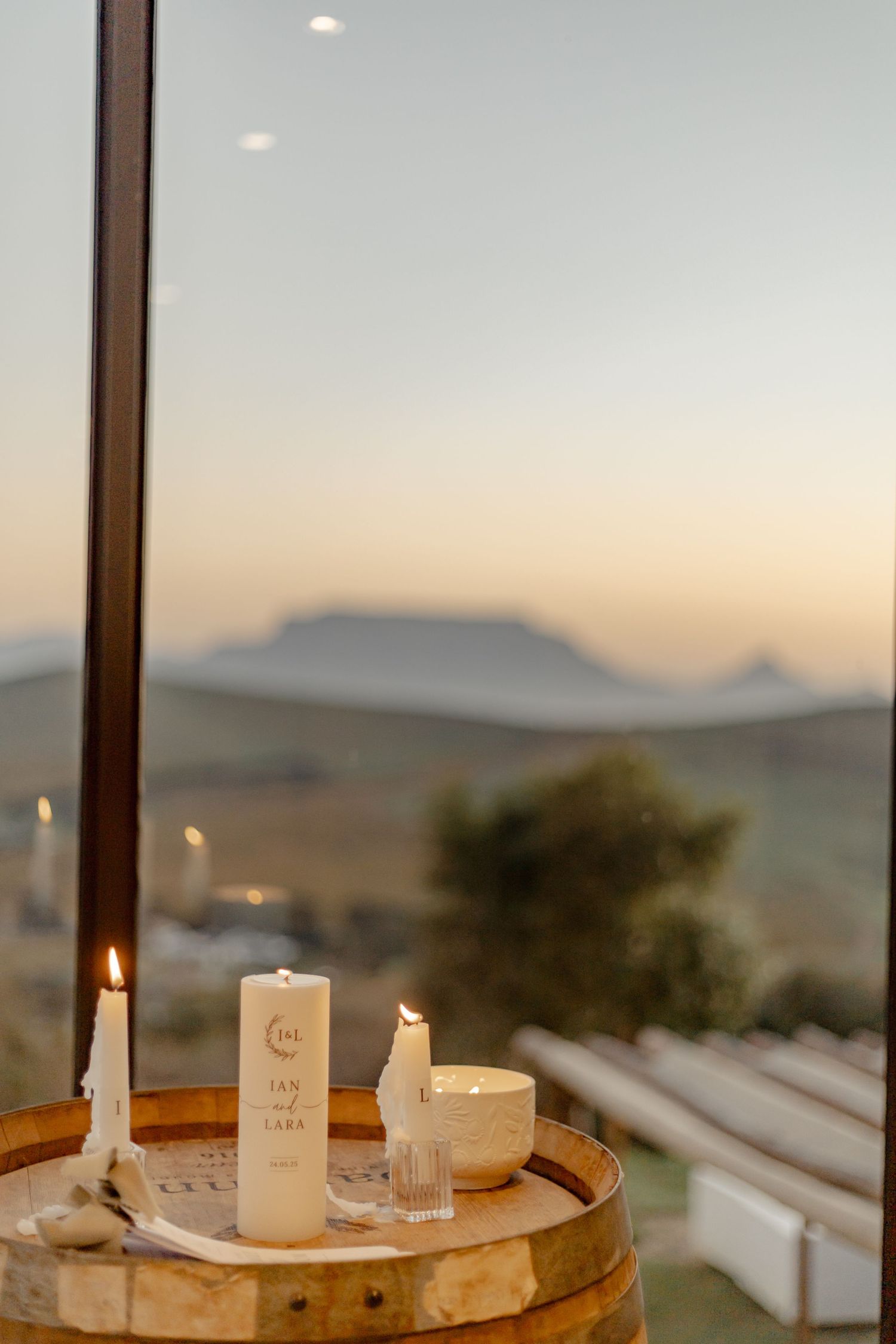 Candles arranged on a wooden barrel at sunset with mountain silhouettes visible through large window panels in background.