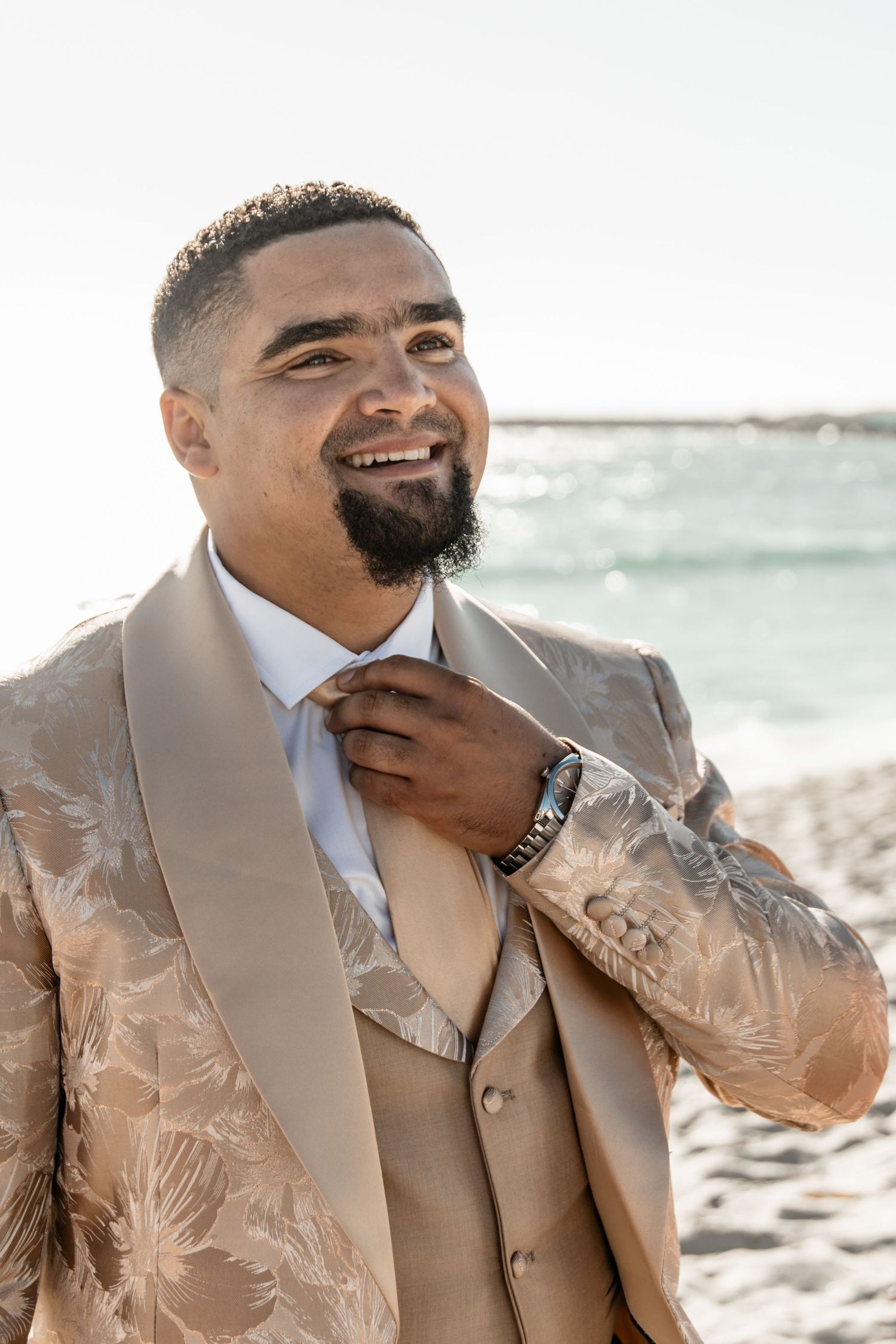 Person wearing a stylish beige floral suit jacket adjusts their tie while smiling at a beach setting.