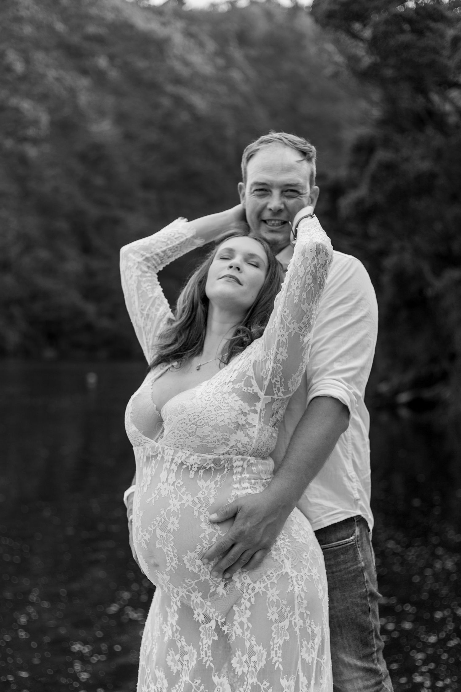 A romantic black and white maternity photo shows a couple embracing outdoors near a lake.