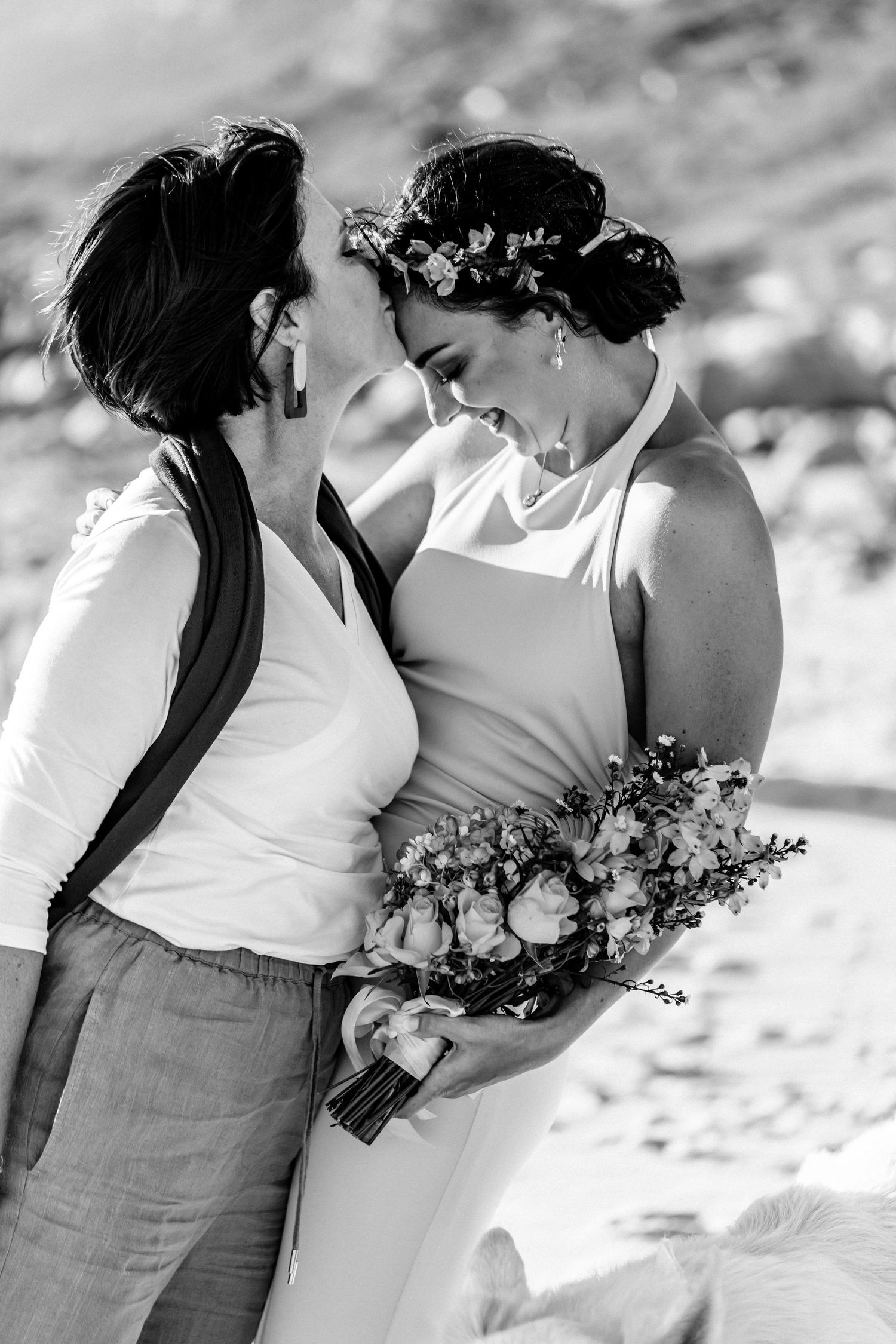 A romantic black and white beach wedding photo showing two brides sharing an intimate moment with a floral bouquet.