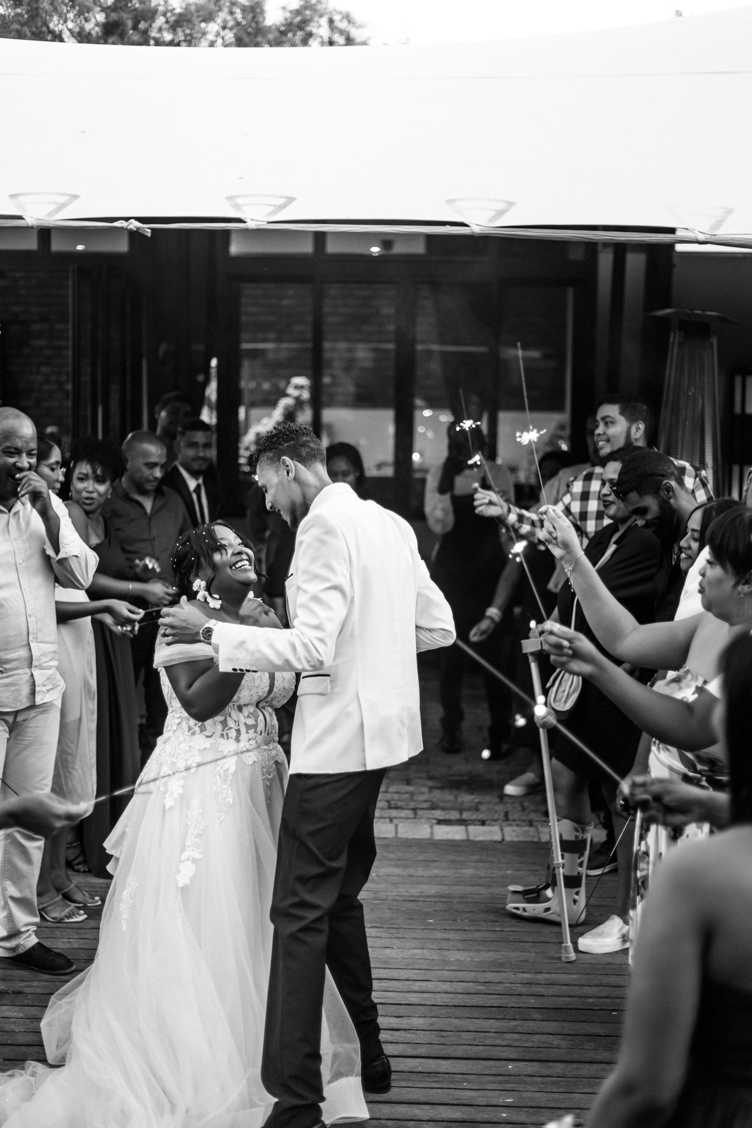 A couple sharing their first dance at a wedding reception on a wooden deck while guests look on and capture photos.