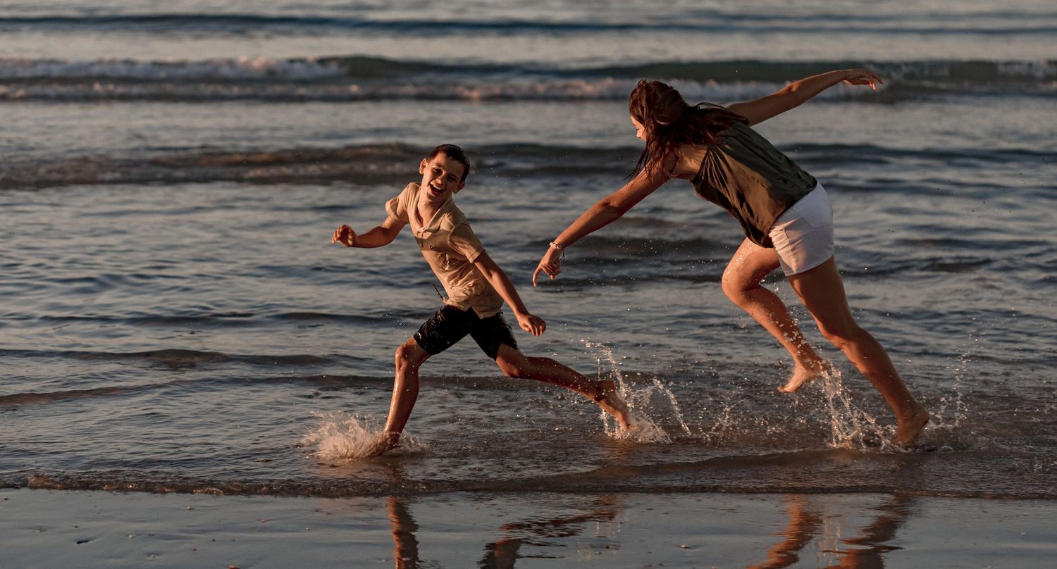 Two people running and splashing through shallow ocean water at sunset along a beach.