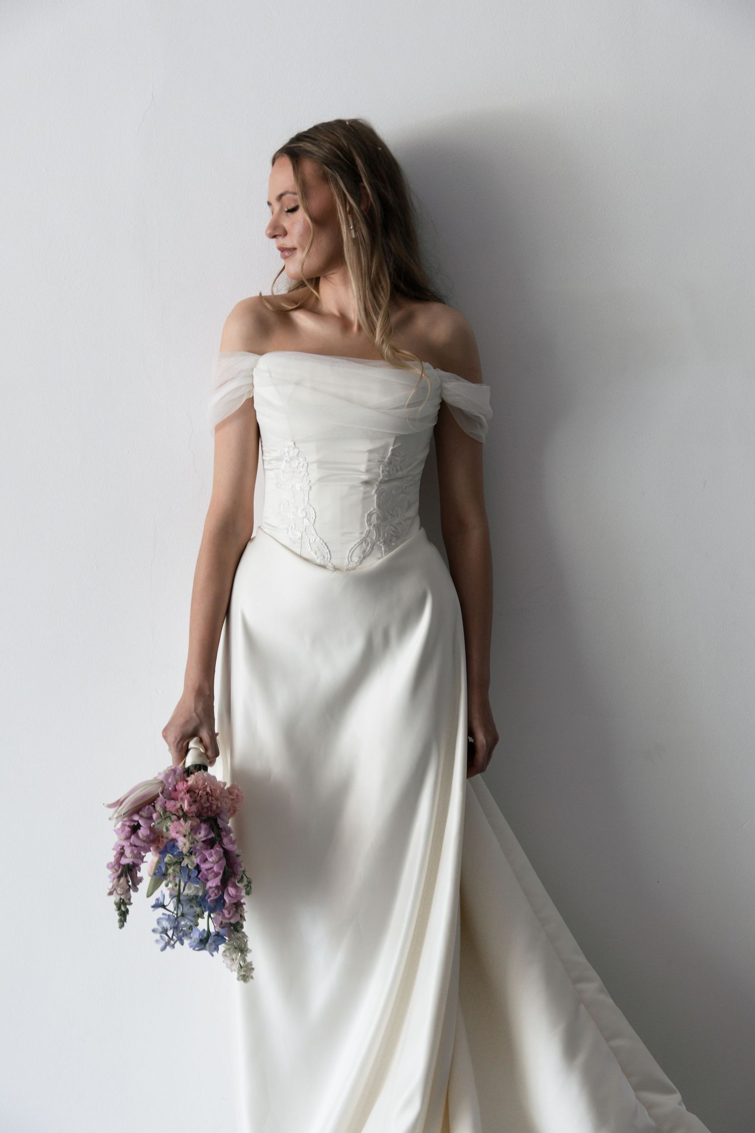 A bride in an elegant off-shoulder white wedding gown poses against a light wall holding a purple and pink bouquet.