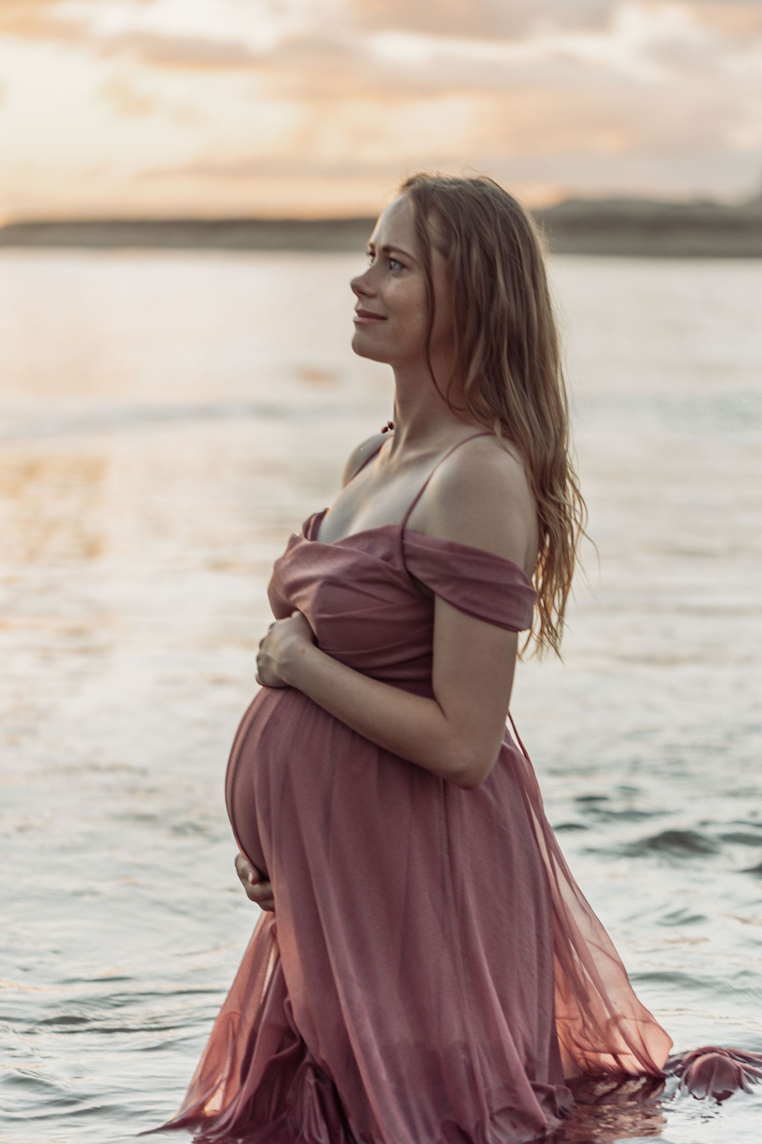 A pregnant woman in a mauve dress stands in water at sunset, cradling her baby bump in a serene maternity photoshoot.