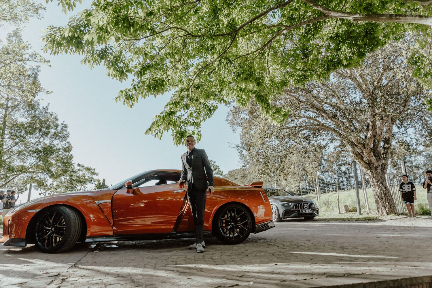 Man standing beside a vibrant orange Nissan GT-R sports car parked under shady trees on a sunny day.