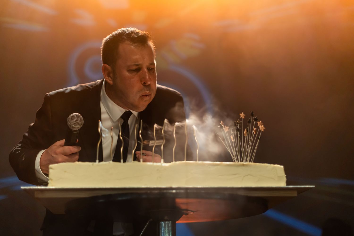 Man in suit blowing out candles on white birthday cake while holding microphone on stage.