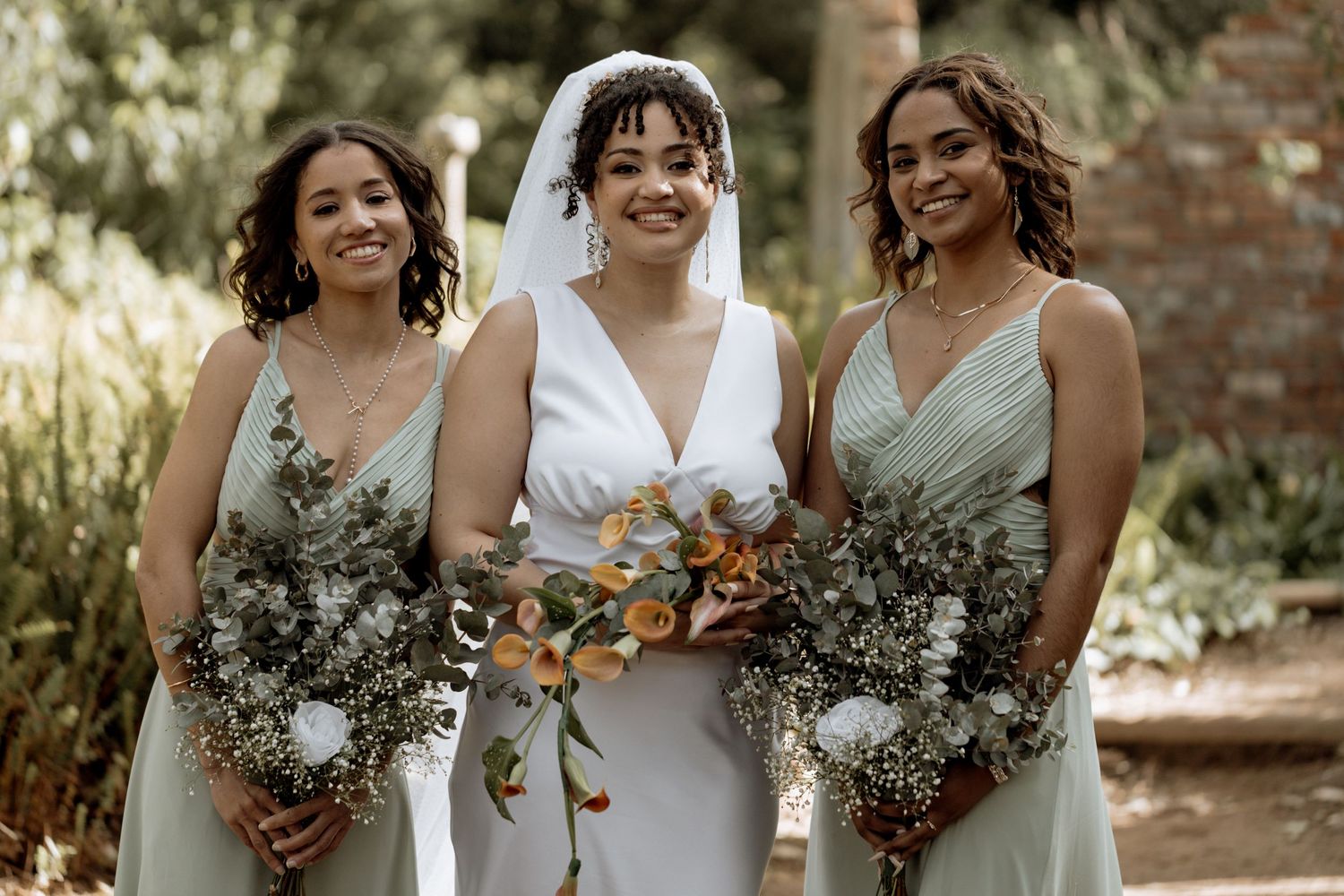 Three women in sage green and white dresses pose together holding beautiful bouquets of mixed flowers and eucalyptus.