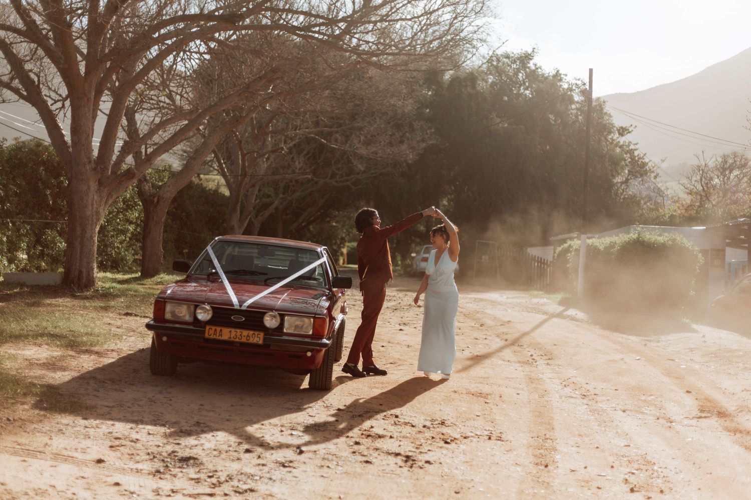 Golden sunlight illuminates a moody desert landscape with a lone red vintage car beneath a barren tree.