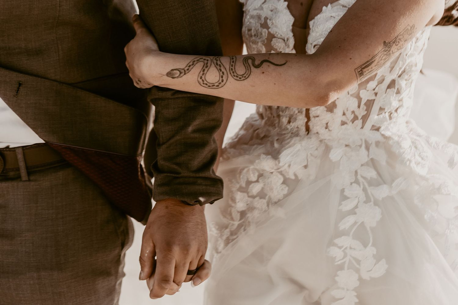 Close-up of a bride and groom holding hands, showcasing a snake tattoo on the groom's forearm and a floral lace wedding dress.