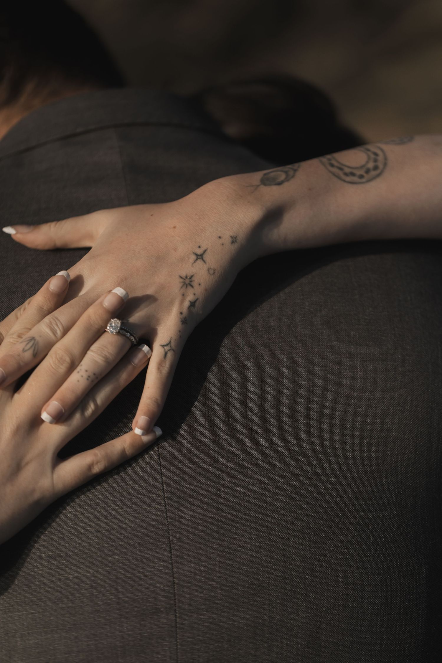 A tattooed hand with a diamond engagement ring rests on a gray suit jacket.