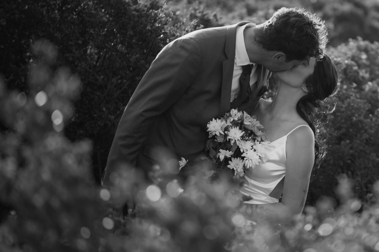 Black and white photo of a couple kissing surrounded by flowers, bride holding bouquet, groom in suit.