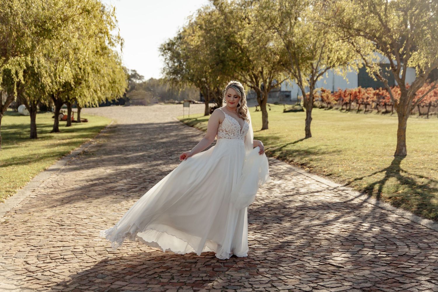 Bride in a white gown walks along a scenic cobblestone path under autumn trees.