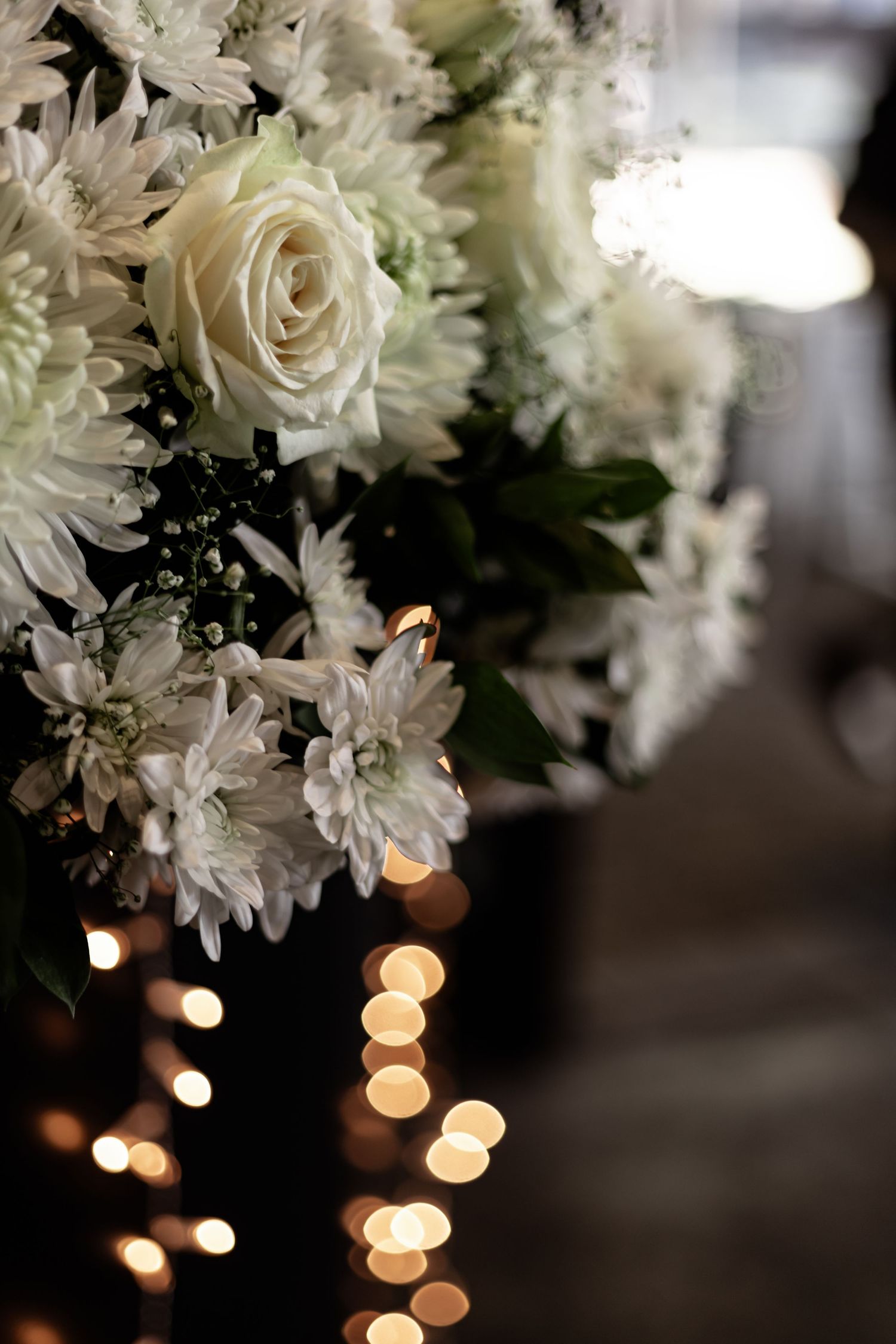 A romantic arrangement of white roses and daisies with soft bokeh lights glowing in the background.