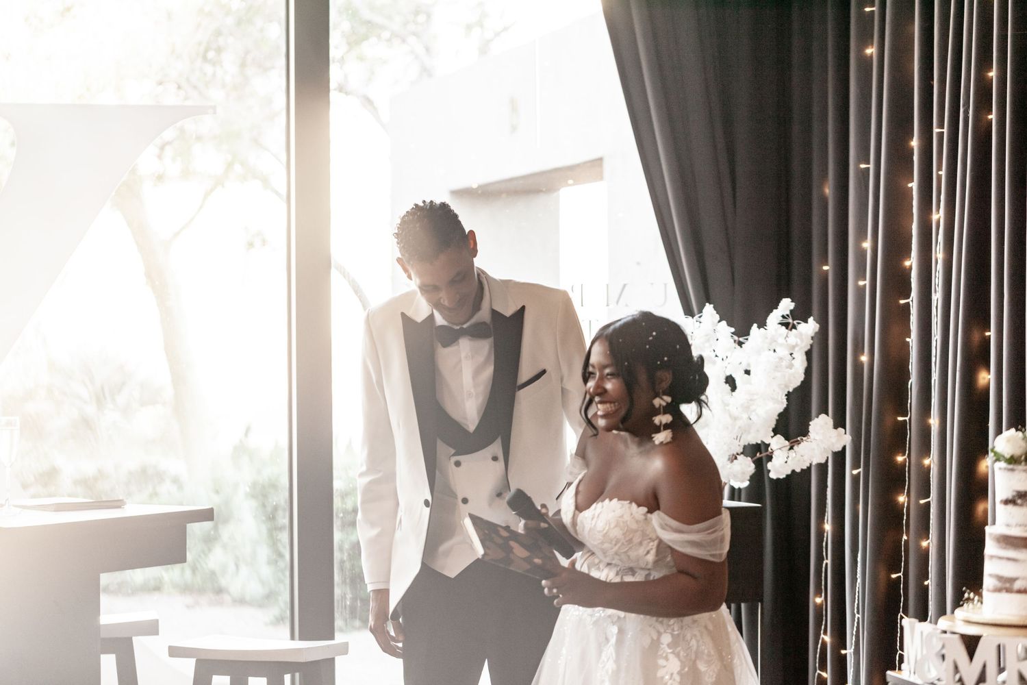 A wedding couple shares a joyful moment together in front of sunlit windows at their reception venue.
