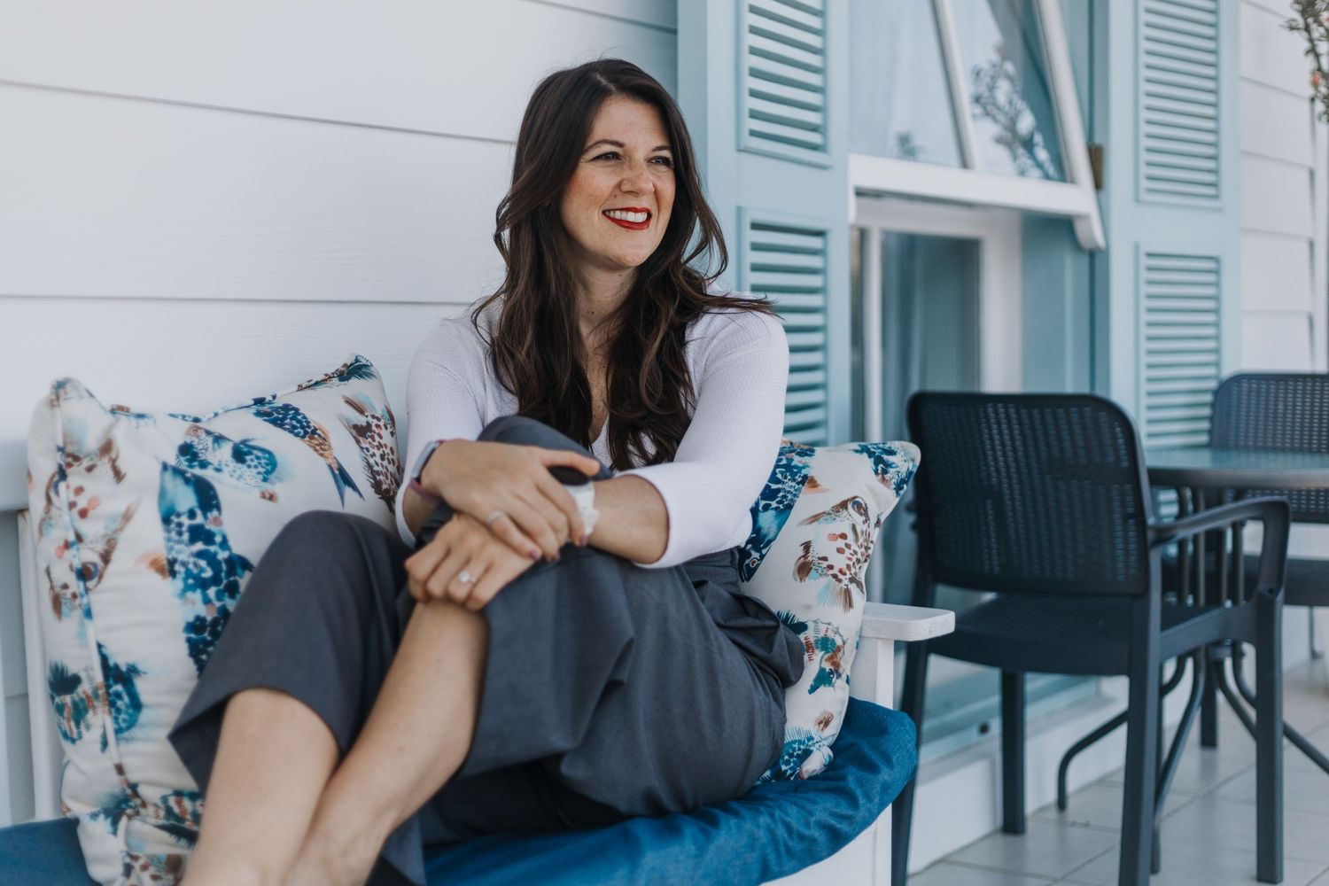 Woman relaxes on floral patterned outdoor patio furniture with light blue shutters in background.