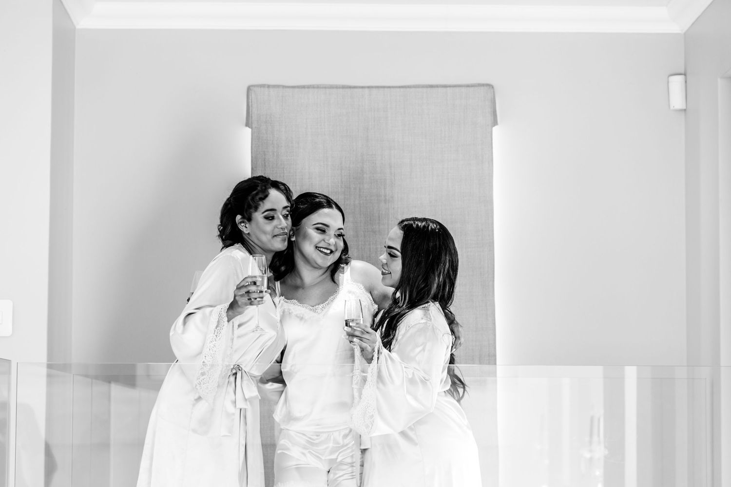 A group in white dresses and veils walk together in a bright hallway during a bridal preparation.