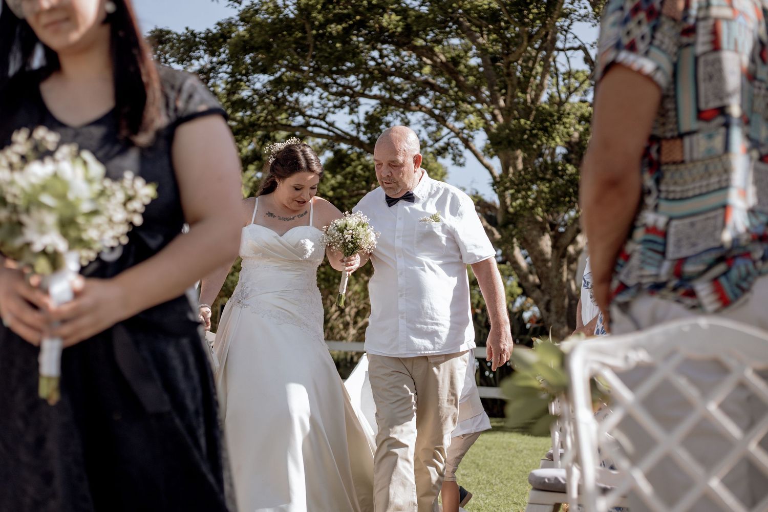 A couple holds hands walking down the aisle at an outdoor wedding ceremony with blooming trees in the background.