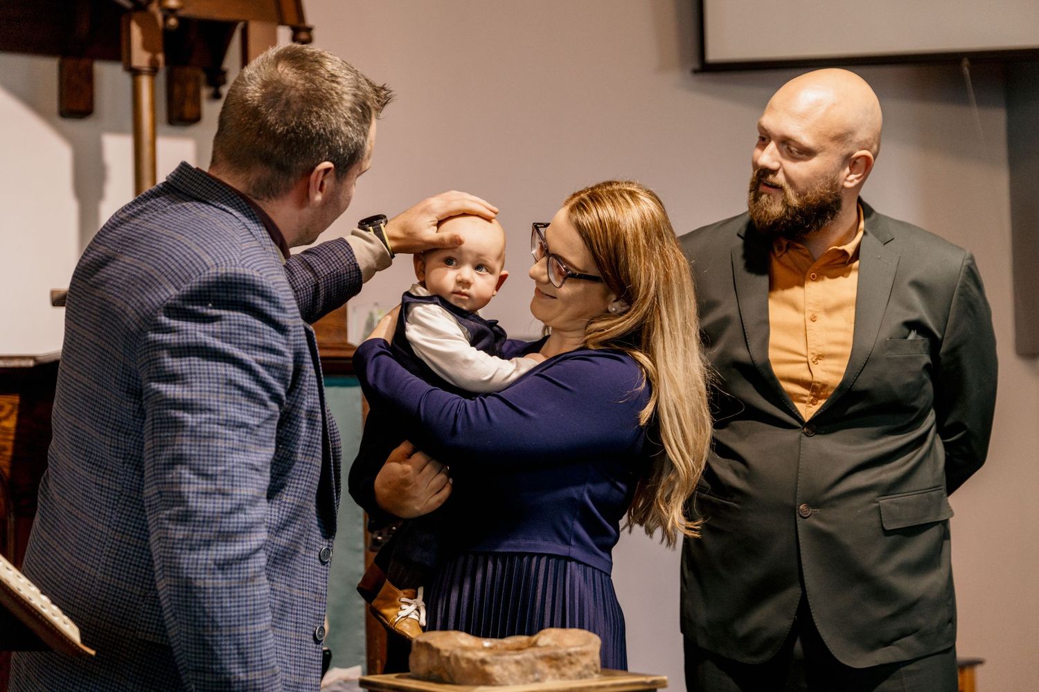 A priest blesses an infant held by a woman while a bald man with a beard watches during a baptism ceremony.