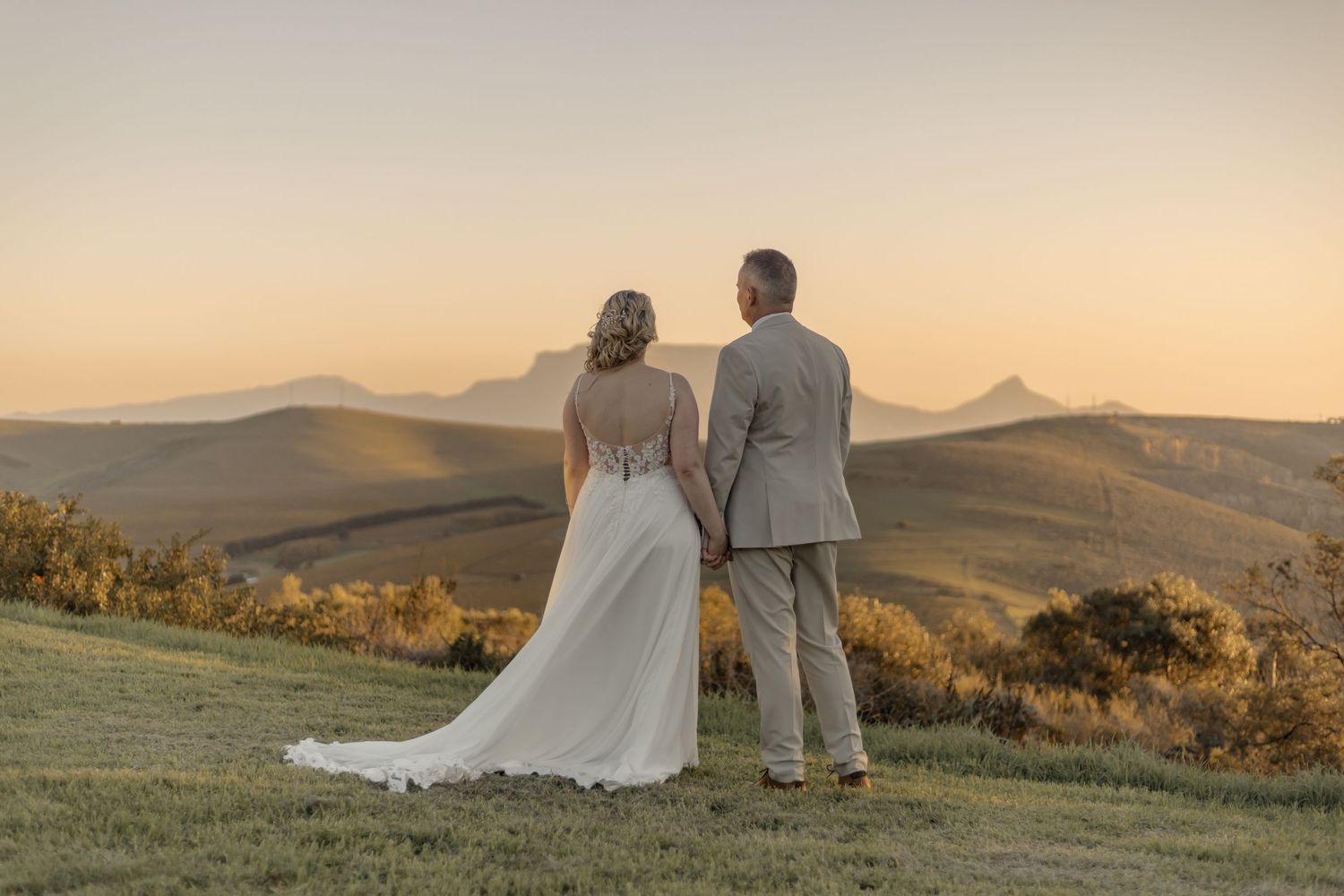 A couple in wedding attire stands together overlooking rolling hills at sunset in a scenic mountain landscape.