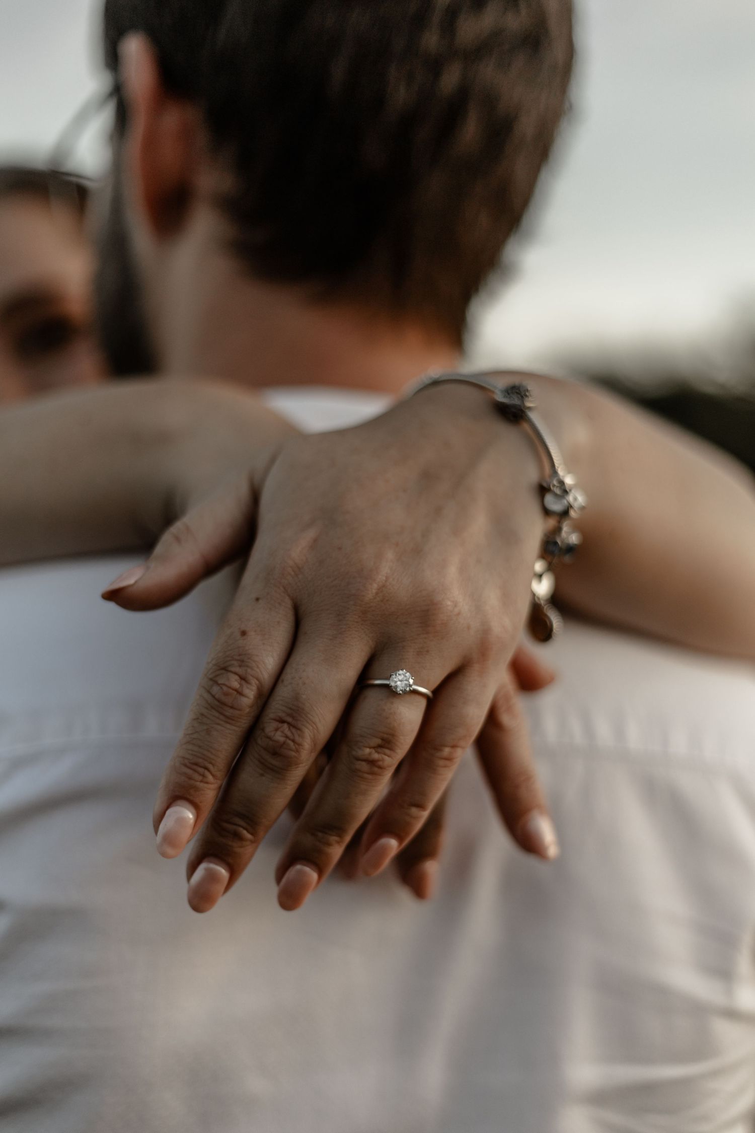 Close-up of a hand wearing an engagement ring and bracelet resting gently on someone's shoulder in soft lighting.