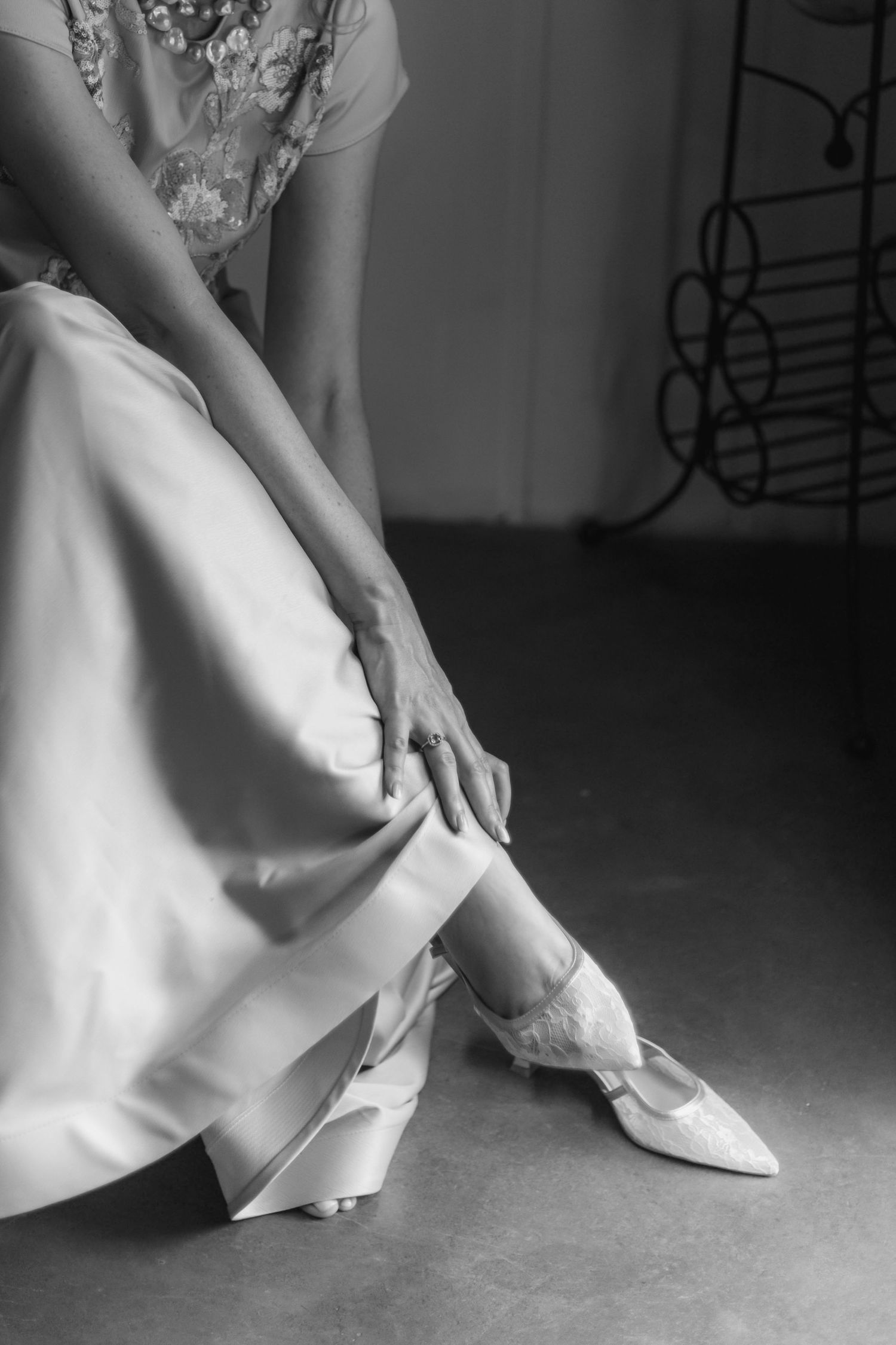 Black and white photo of someone adjusting elegant white bridal shoes while wearing a flowing white dress.
