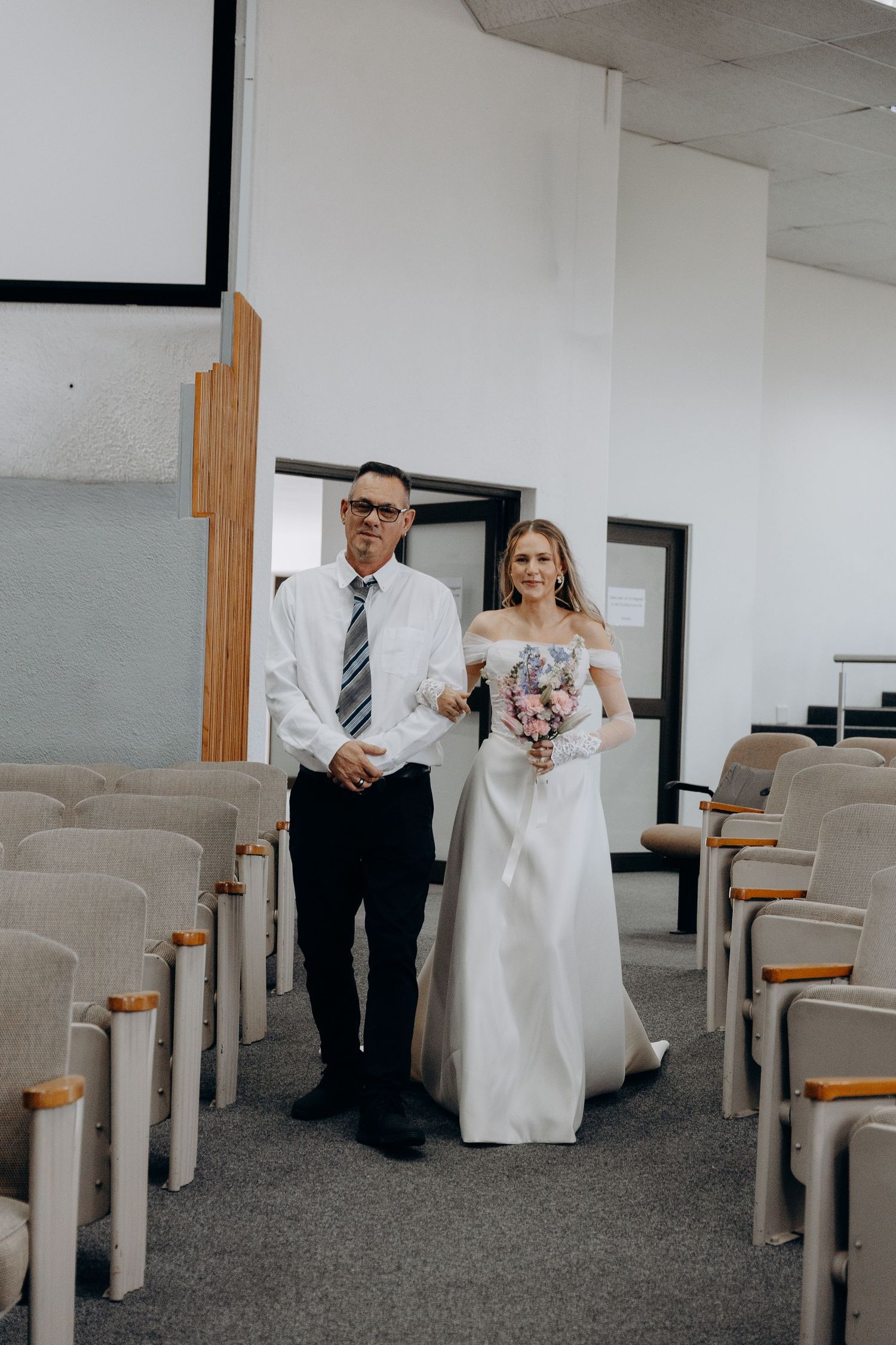 A wedding ceremony processional in a simple church hall with grey carpeting and wooden pews.