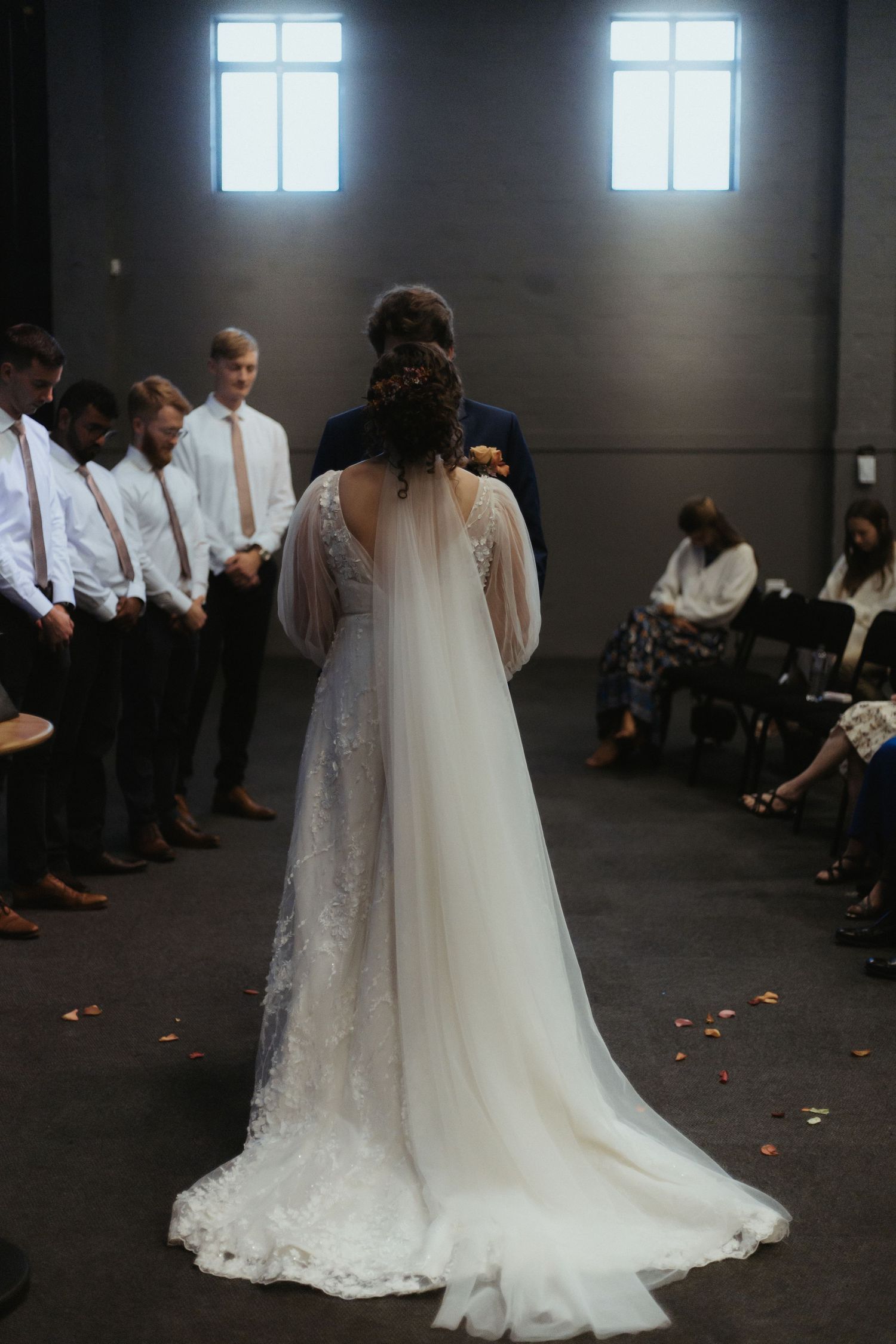 A wedding ceremony scene with white floral petals scattered on dark floor as a bride in flowing gown walks past seated guests.