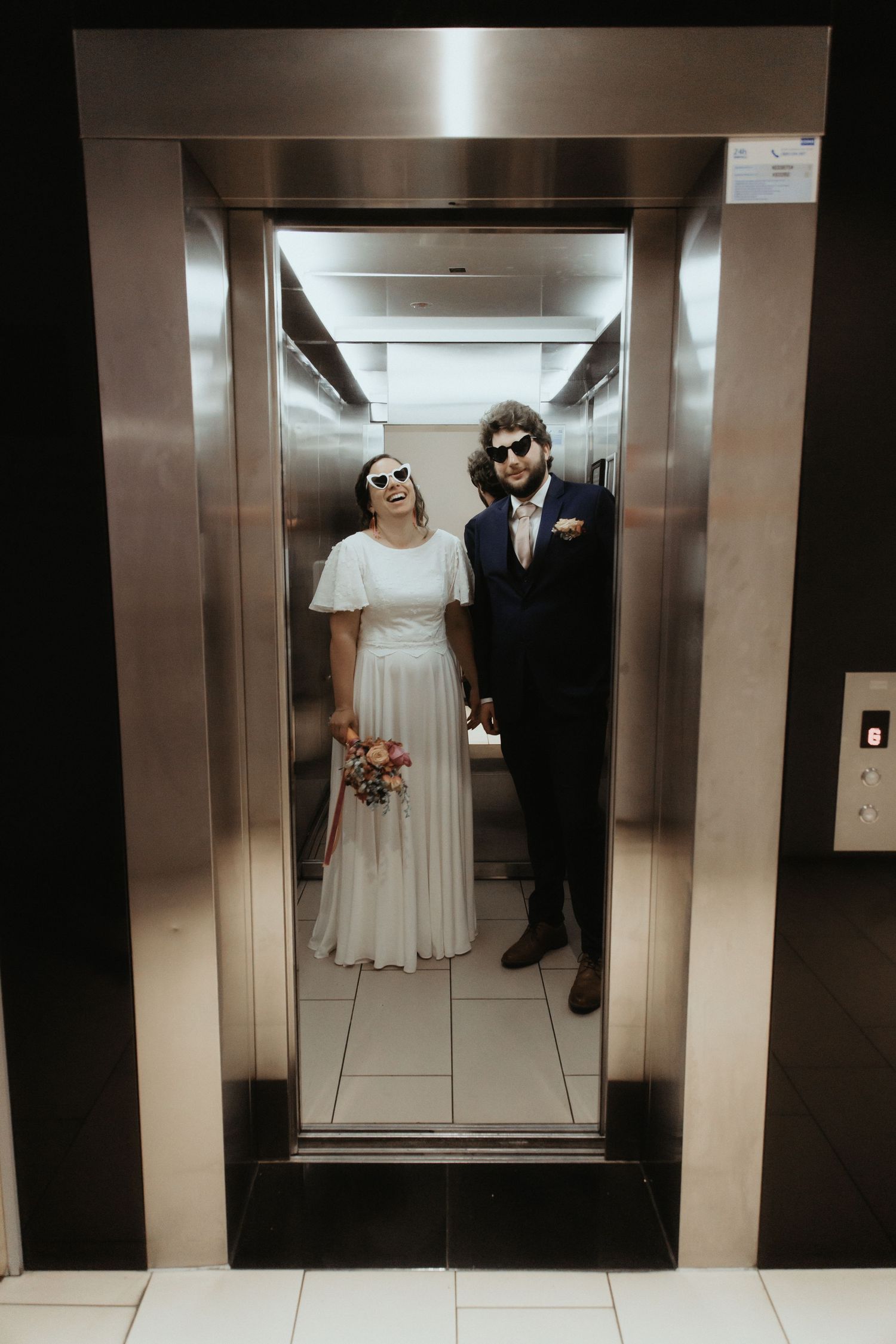 A couple in wedding attire poses for artistic shots inside a modern elevator with metallic walls.