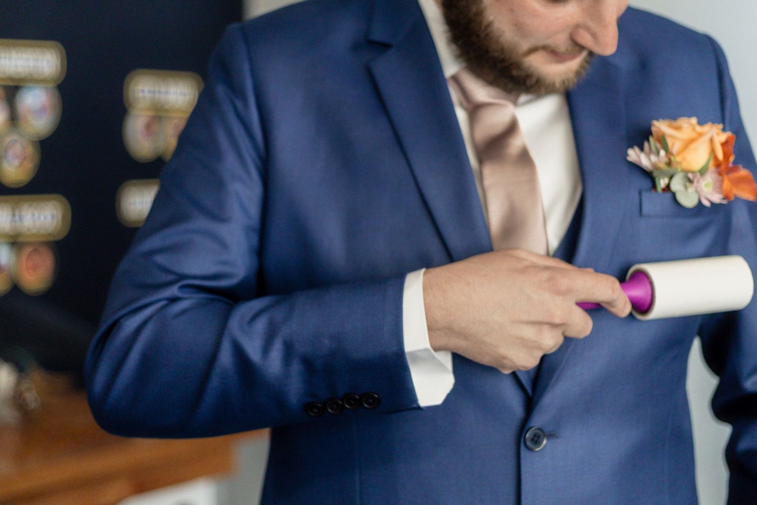 Someone in a navy blue suit with a peach rose boutonniere uses a lint roller on their jacket.