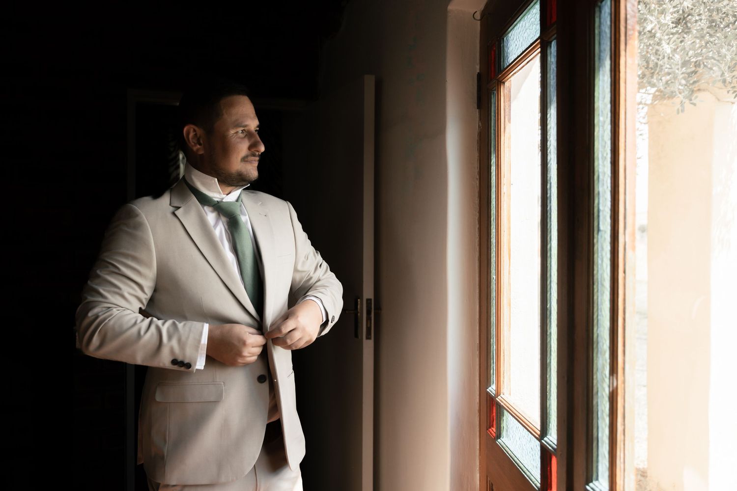 Elegant groom in a beige suit adjusting his jacket while standing by a sunlit window.